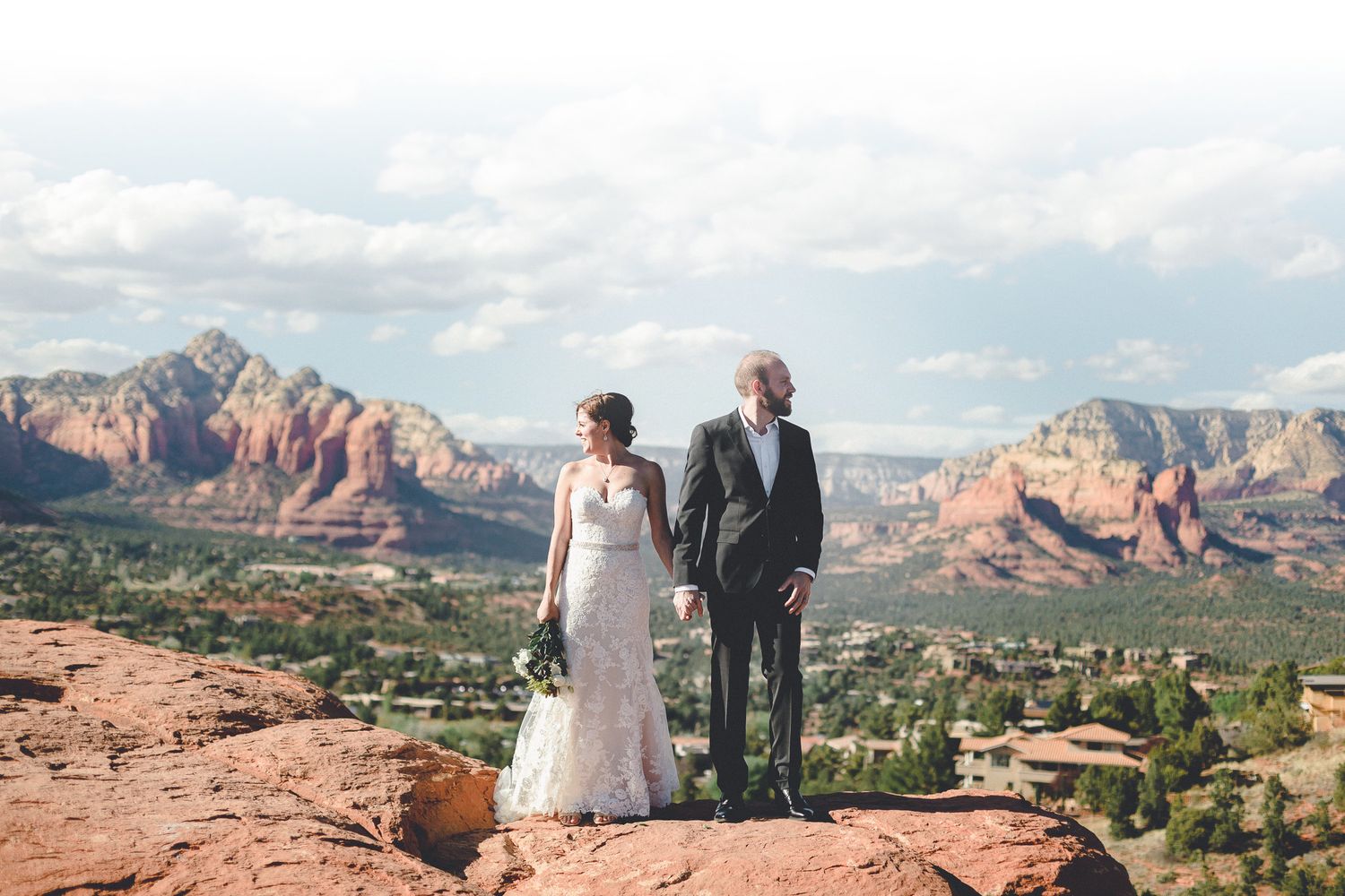 Bride and groom holding hands on red rock overlook with Sedona Arizona landscape behind.