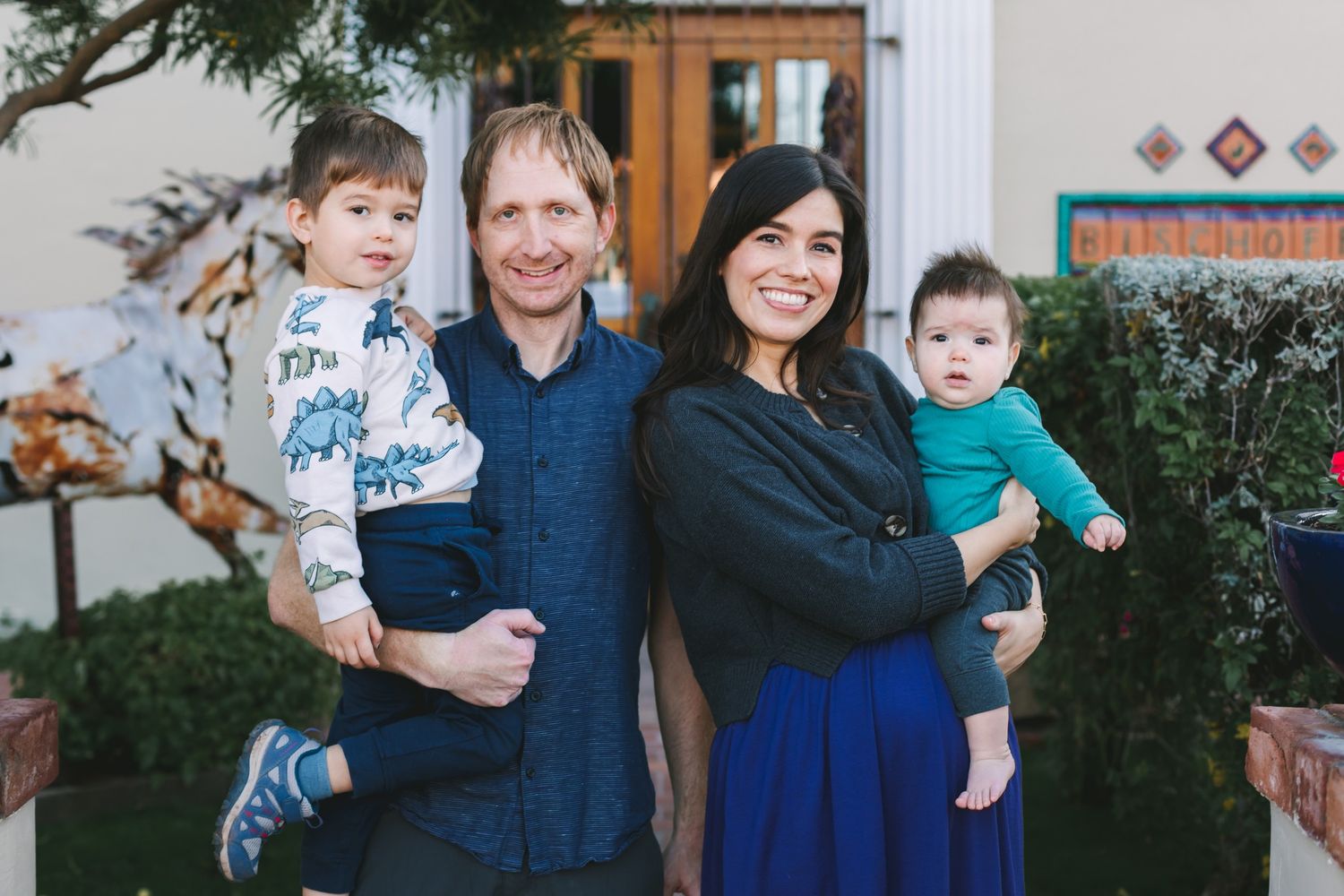 Young family of four posing outdoors, parents holding two small children near a colorful doorway.