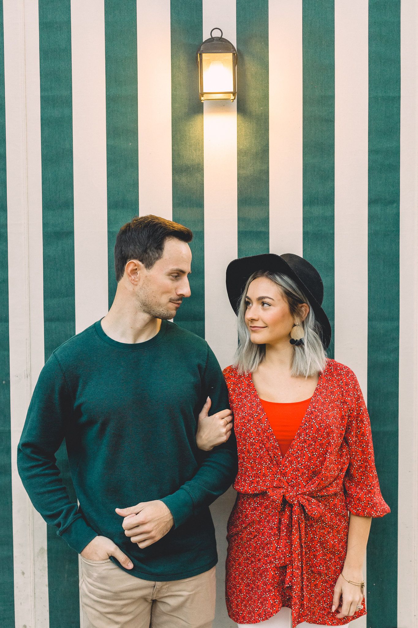 Young couple standing together against a green and white striped wall under a vintage lantern.
