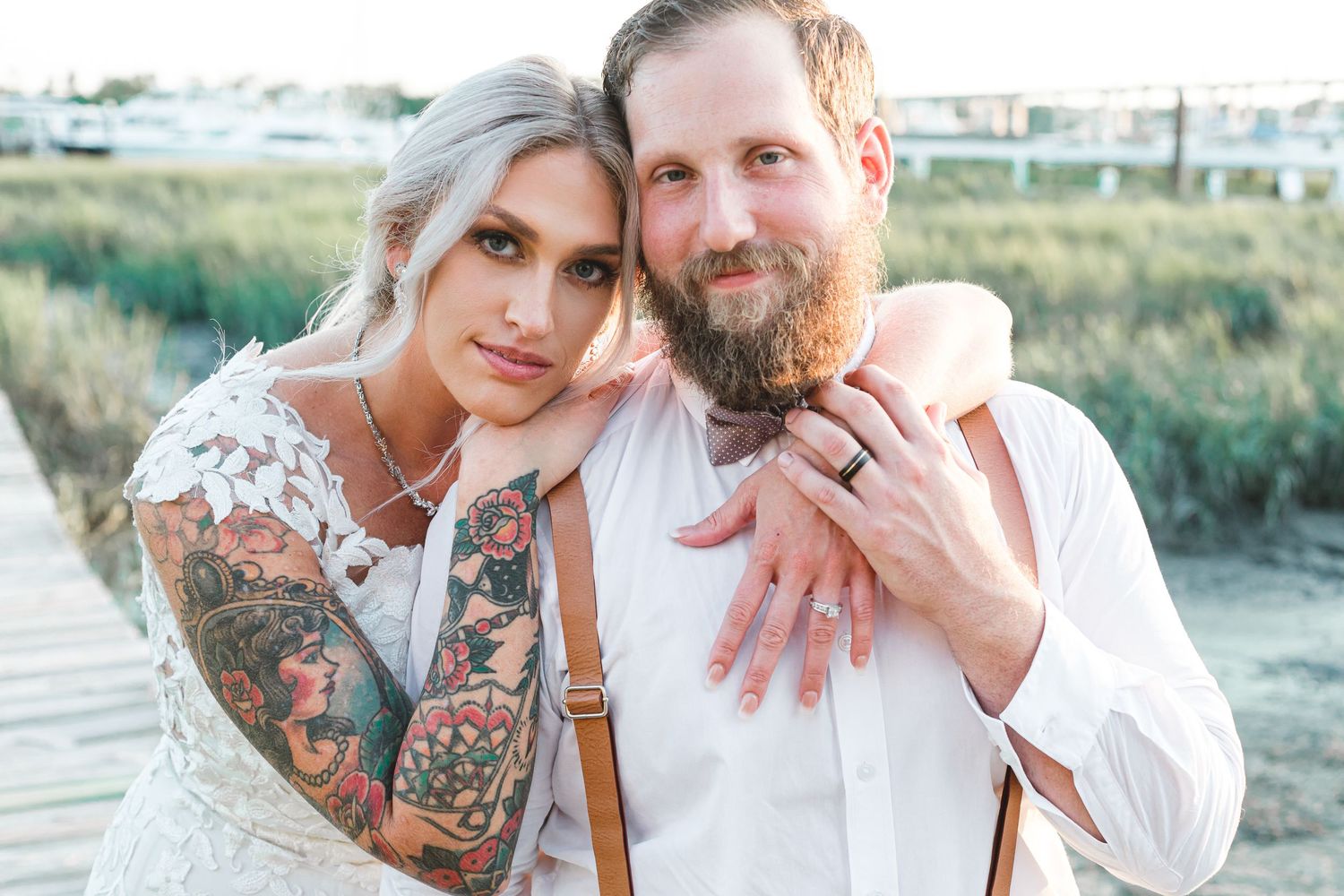Tattooed bride in lace gown embracing bearded groom in white shirt with suspenders at outdoor coastal wedding.