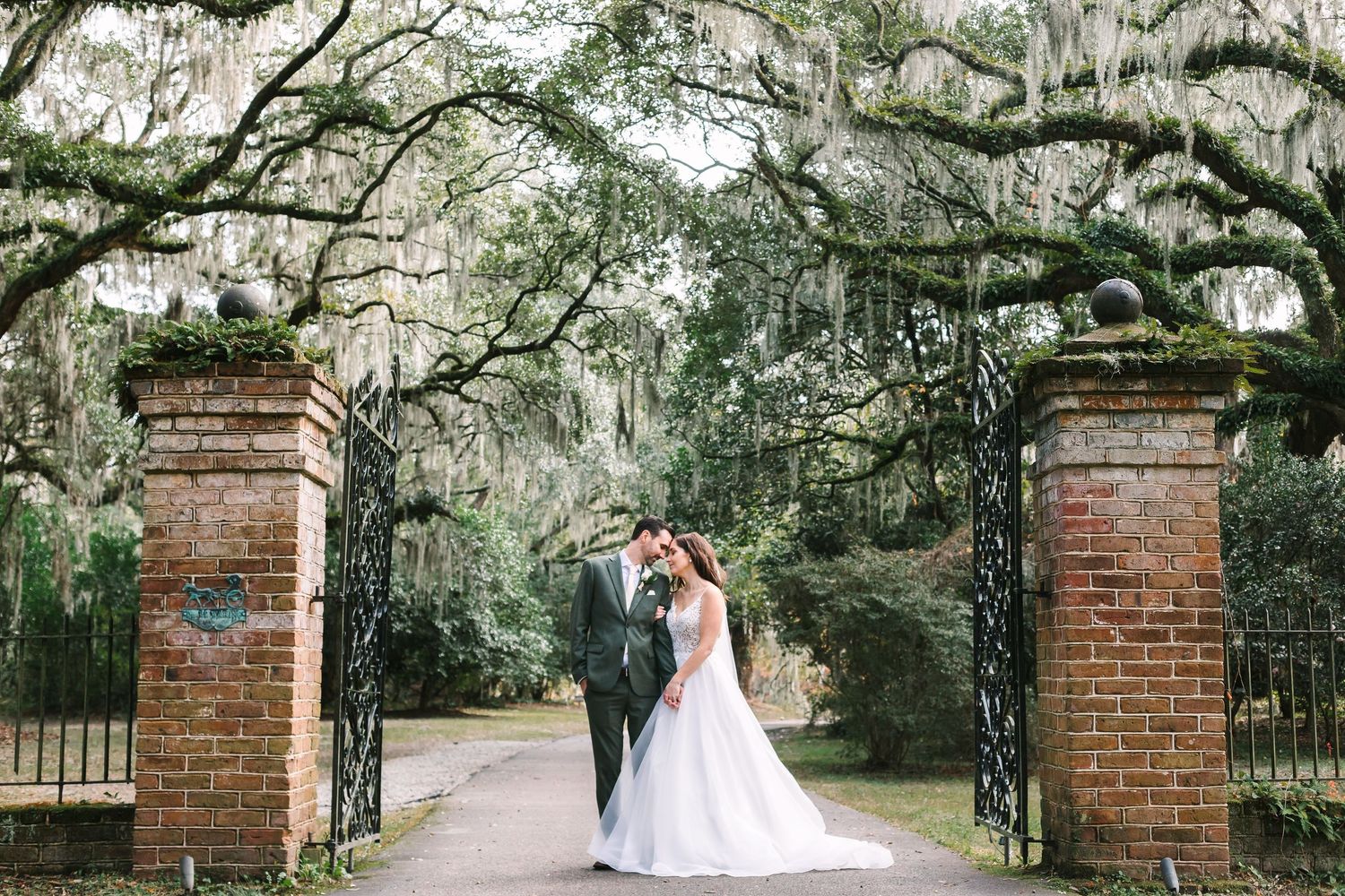 Bride and groom sharing a romantic moment under moss-draped trees framed by ornate iron garden gates.