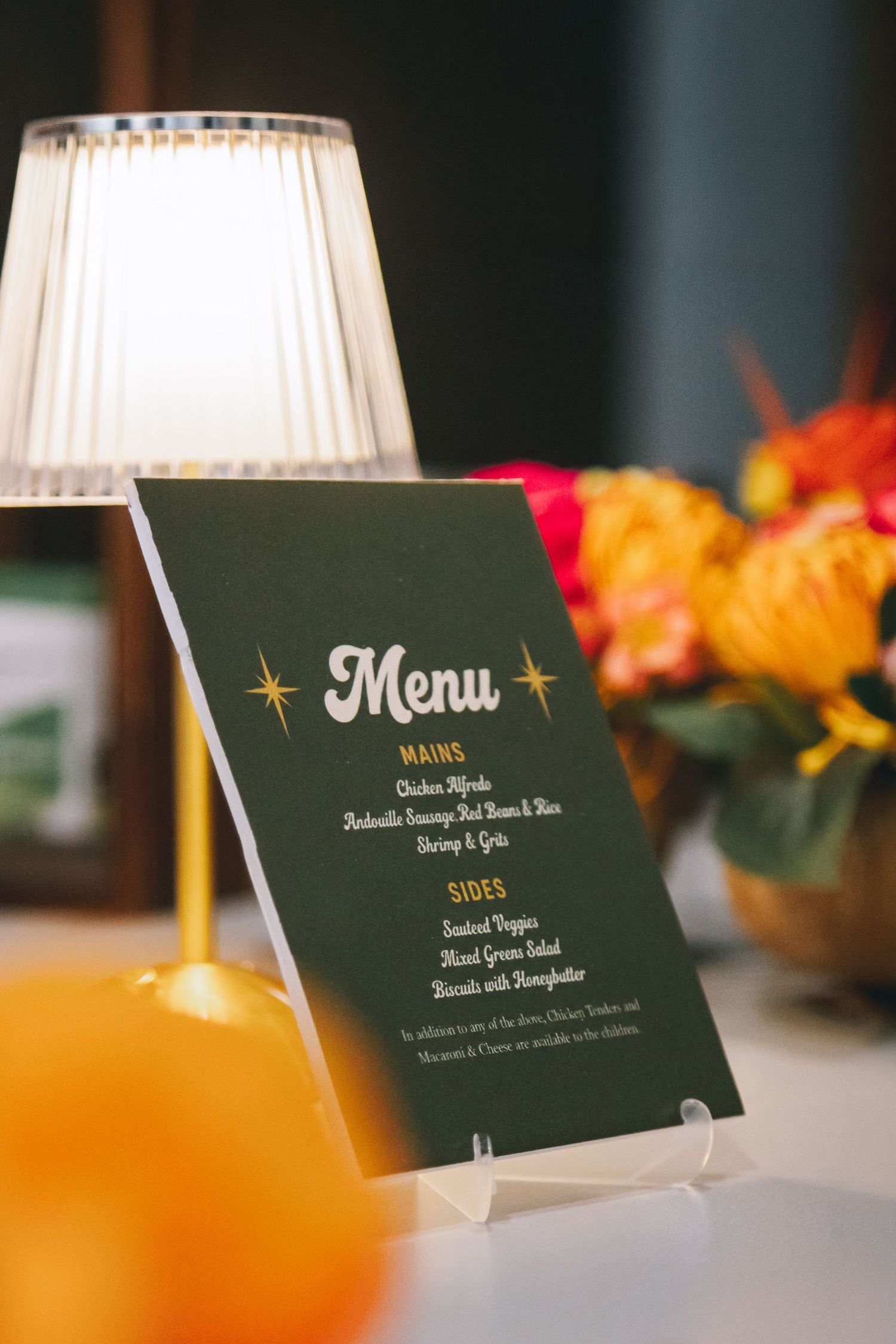 A dark green wedding menu card with gold text displayed on an easel beside a glowing lamp and floral arrangement.