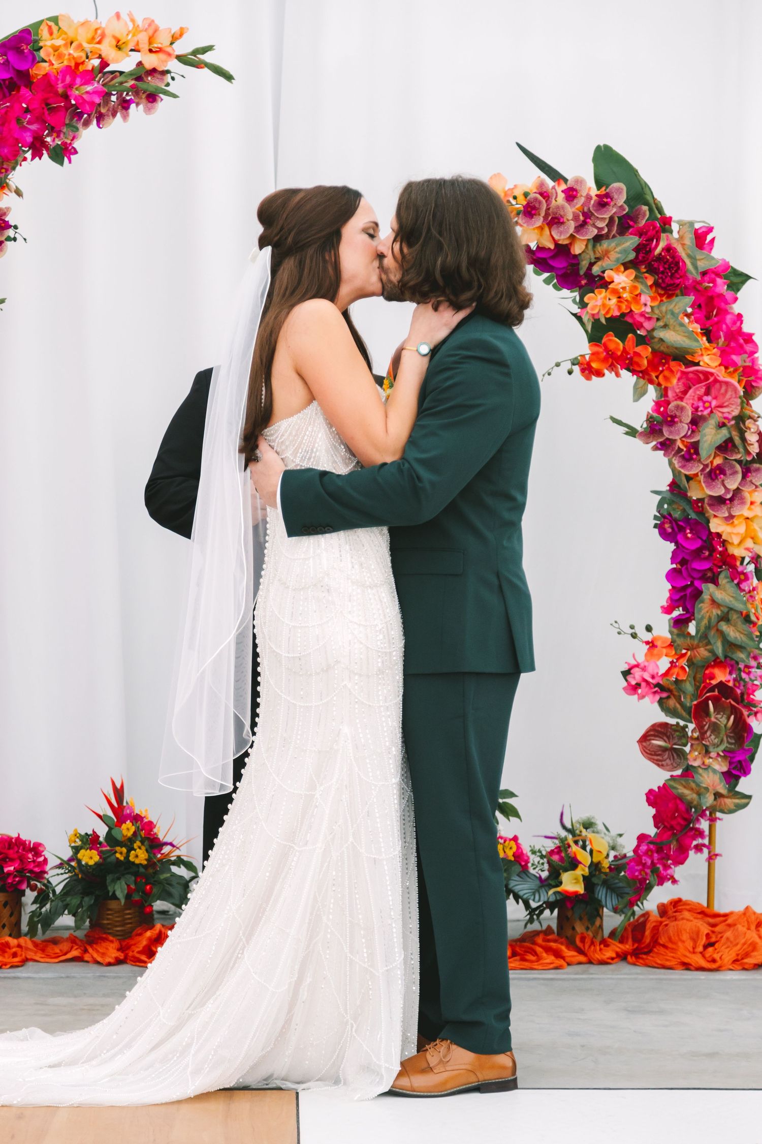 Bride and groom share first kiss at altar adorned with vibrant pink and orange floral arch.