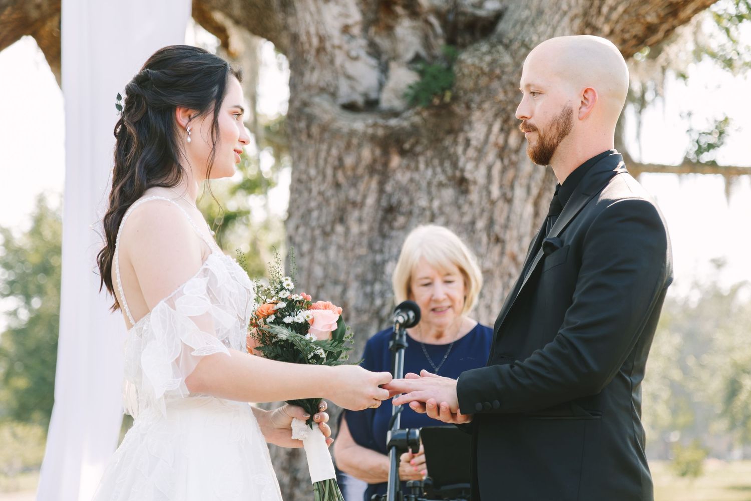 Bride and groom exchange rings during outdoor wedding ceremony beneath large oak tree with officiant.