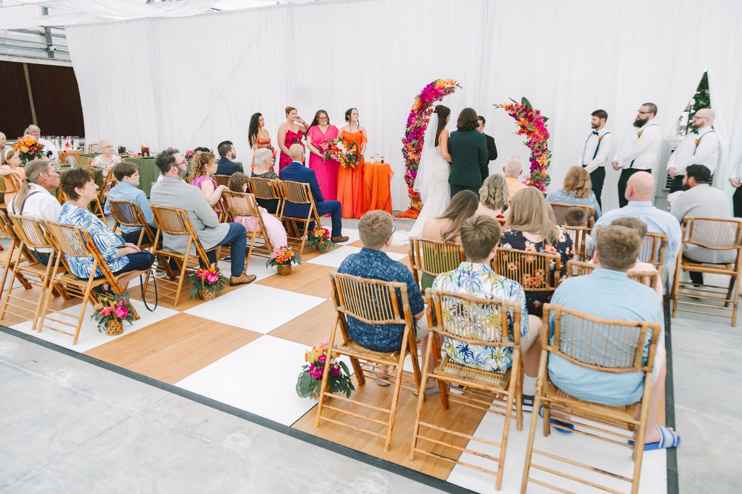 Wedding ceremony with guests seated on wooden chairs facing couple at colorful floral arch.