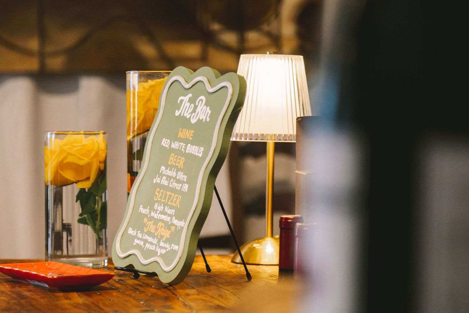 Decorative bar menu sign with yellow flowers and warm lamp lighting at a wedding reception.