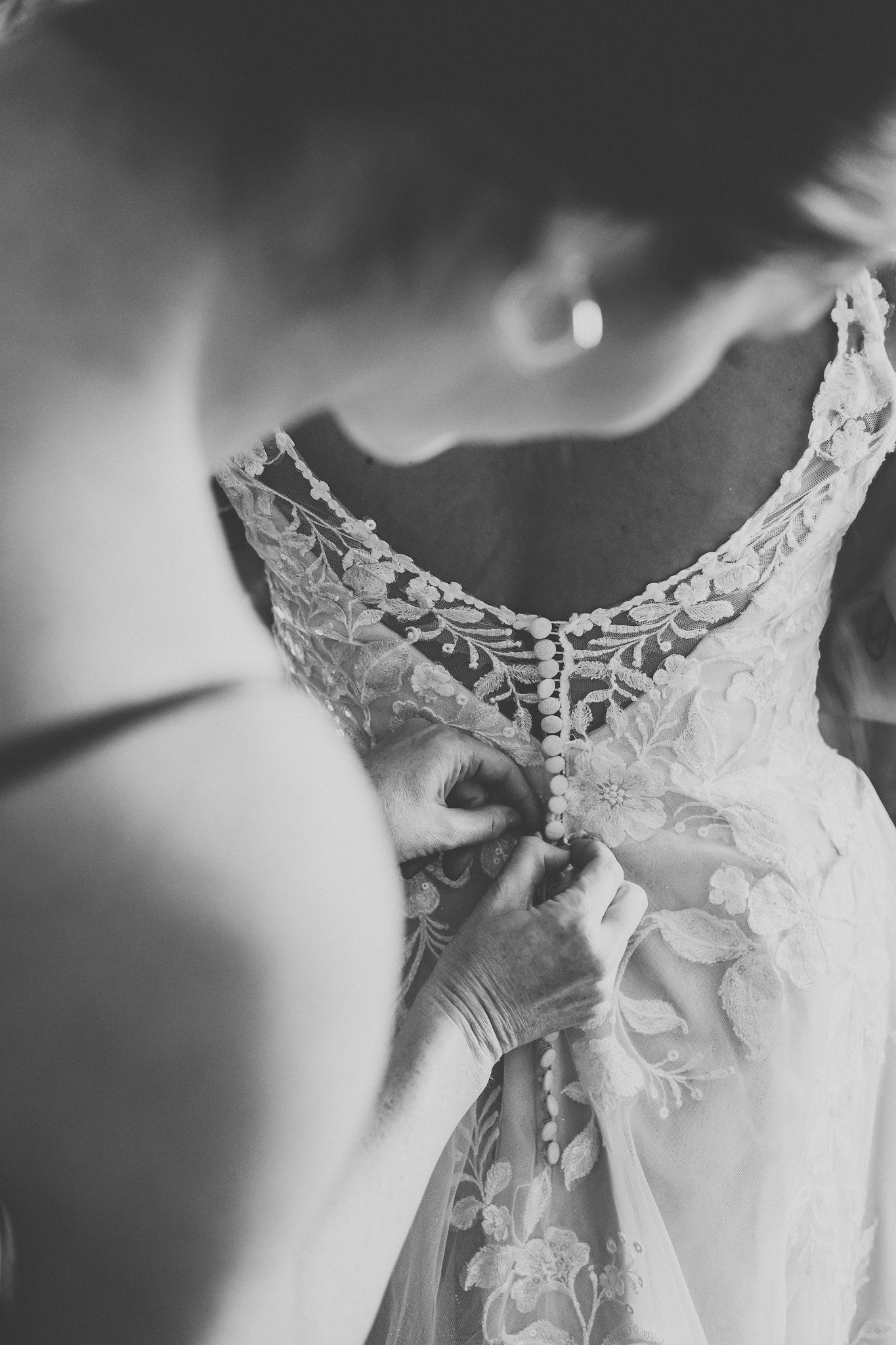 Black and white close-up of bridesmaid carefully fastening delicate buttons on an intricate lace wedding gown.
