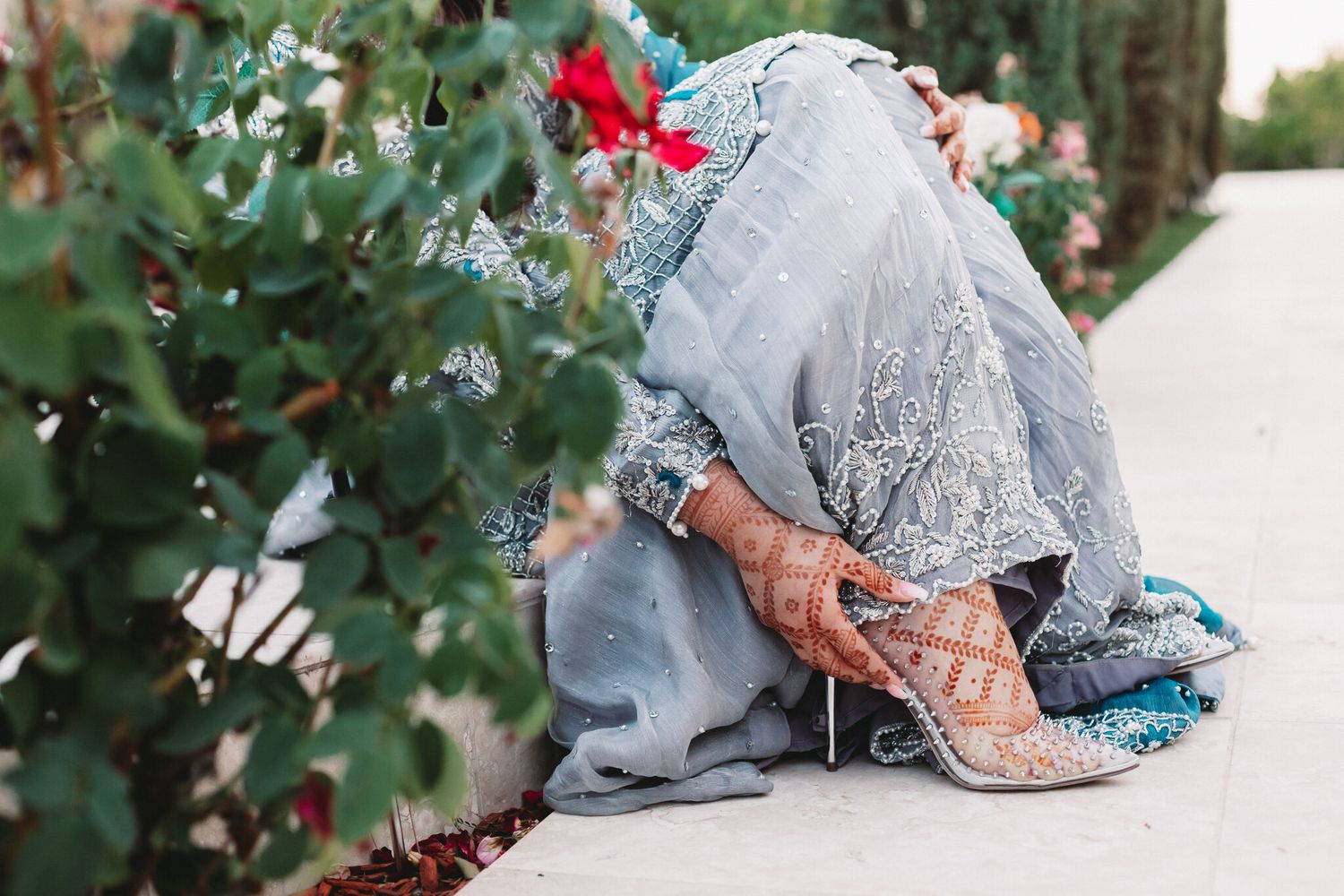 Close-up of a South Asian bride in a blue embroidered lehenga with intricate henna and silver heels near rose bushes.