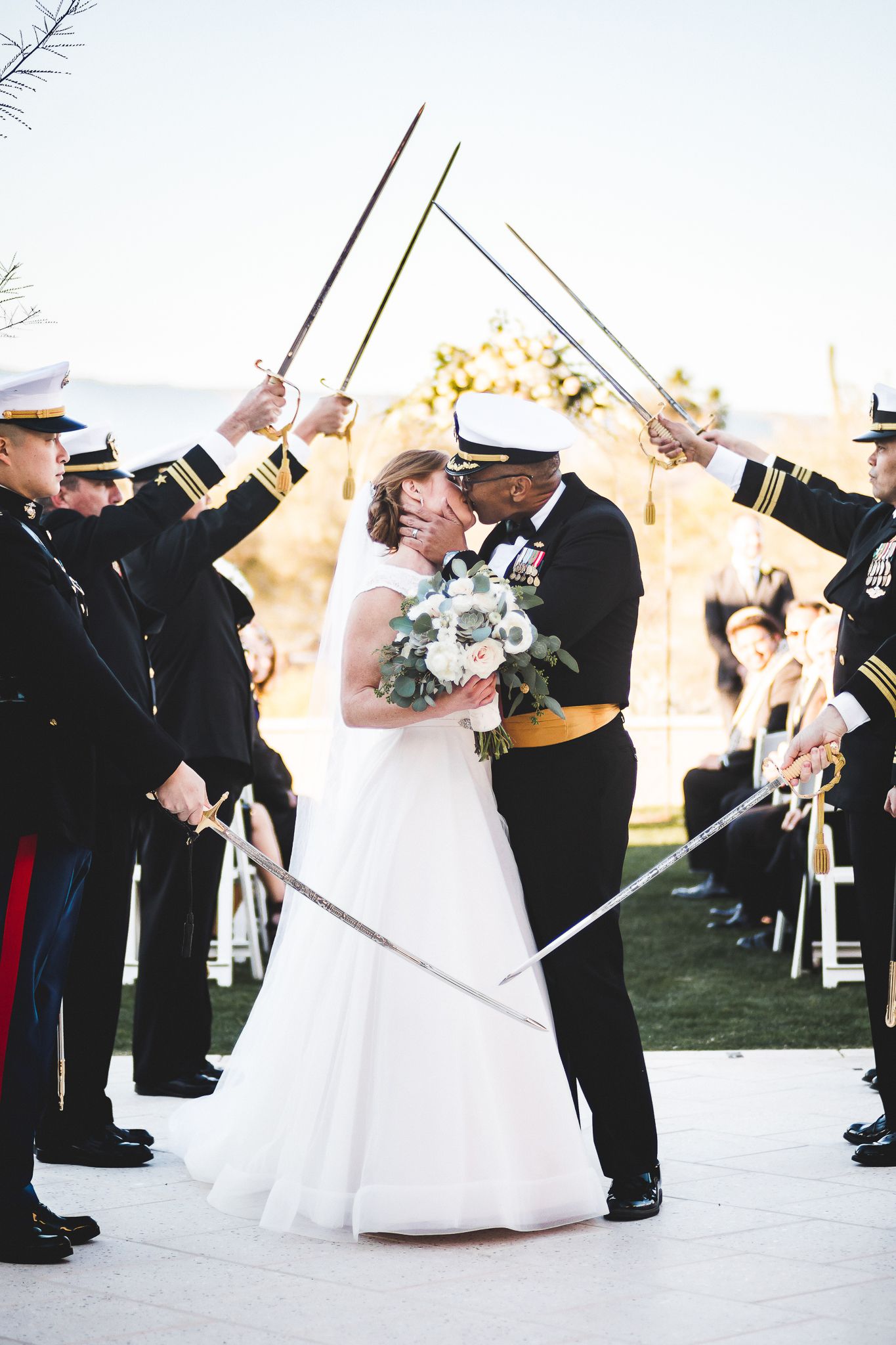 Military wedding couple kissing under a saber arch surrounded by uniformed officers outdoors.