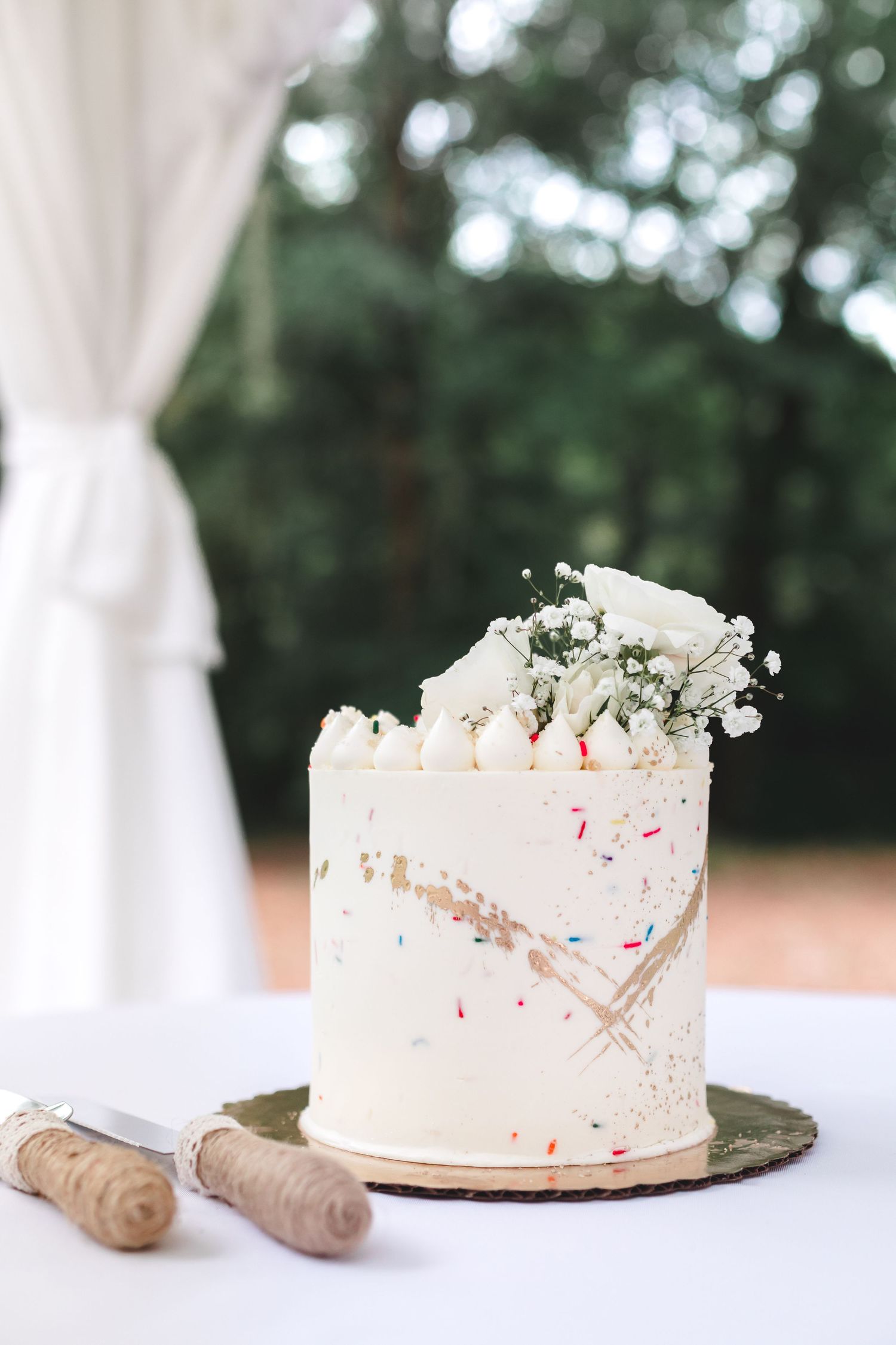 Elegant white wedding cake with colorful sprinkles and fresh white flowers on a rustic slate board outdoors.