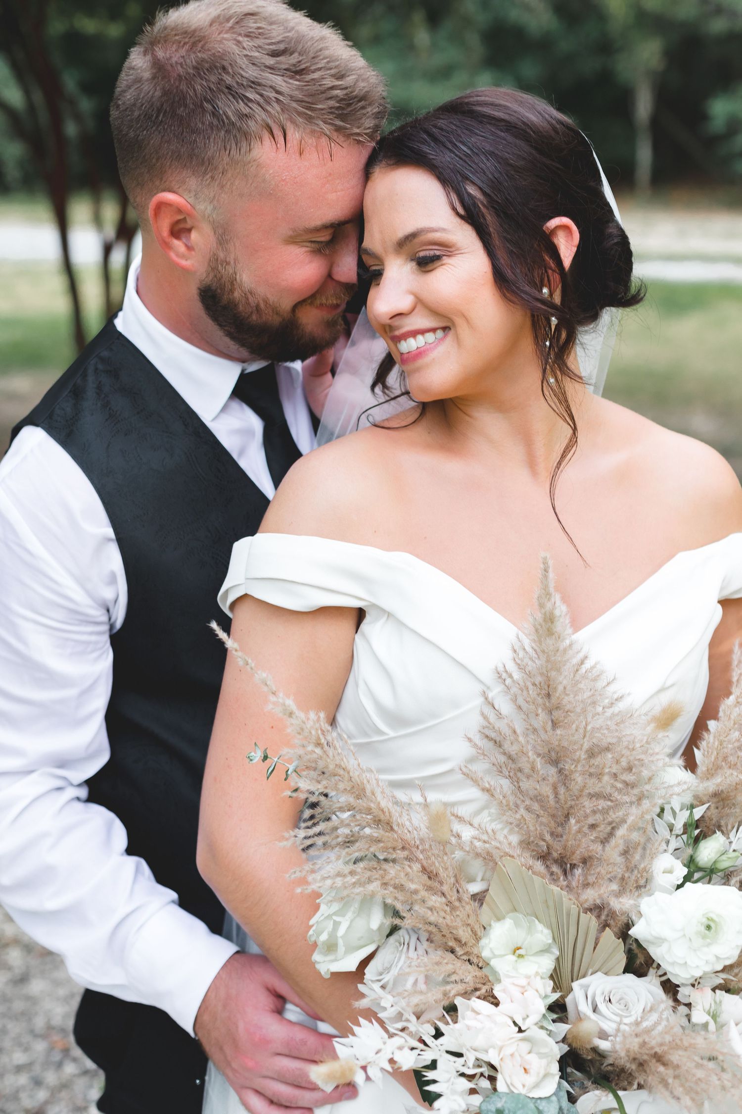 Smiling bride in off-shoulder gown holds pampas grass bouquet while groom tenderly embraces her outdoors.