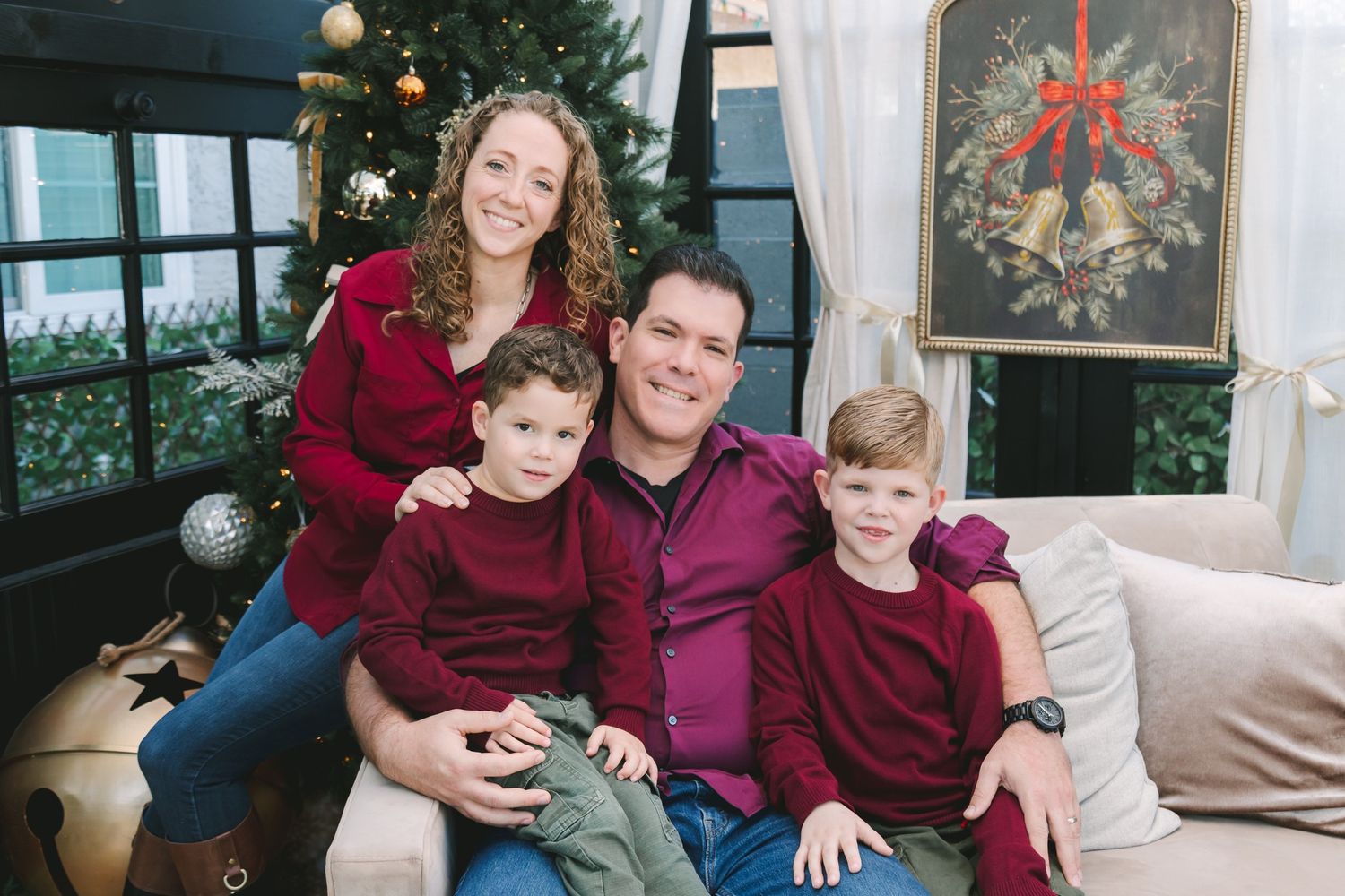 Family of four in red holiday outfits smiling together on a couch with Christmas tree decorations.