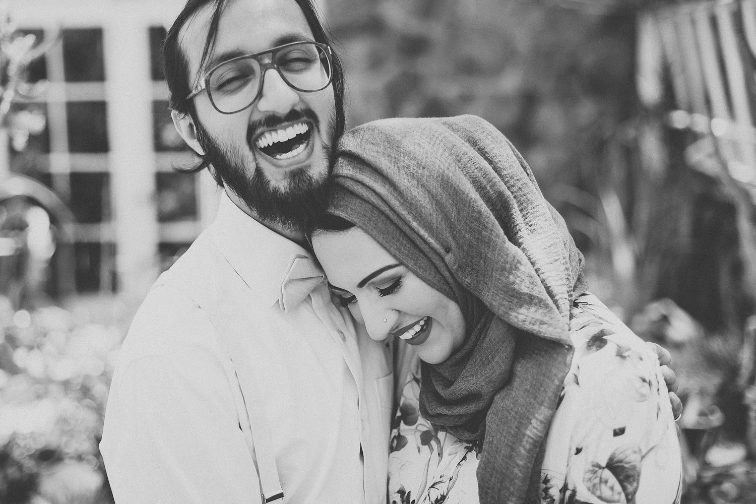 Black and white photo of a laughing couple embracing outdoors, woman wearing a hijab and floral dress.