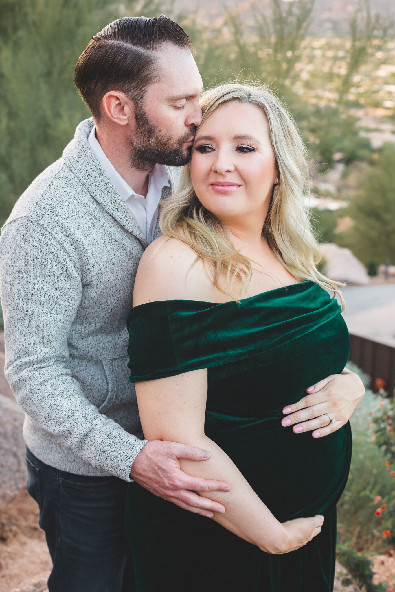 Pregnant woman in green velvet off-shoulder dress being embraced and kissed by her partner outdoors.