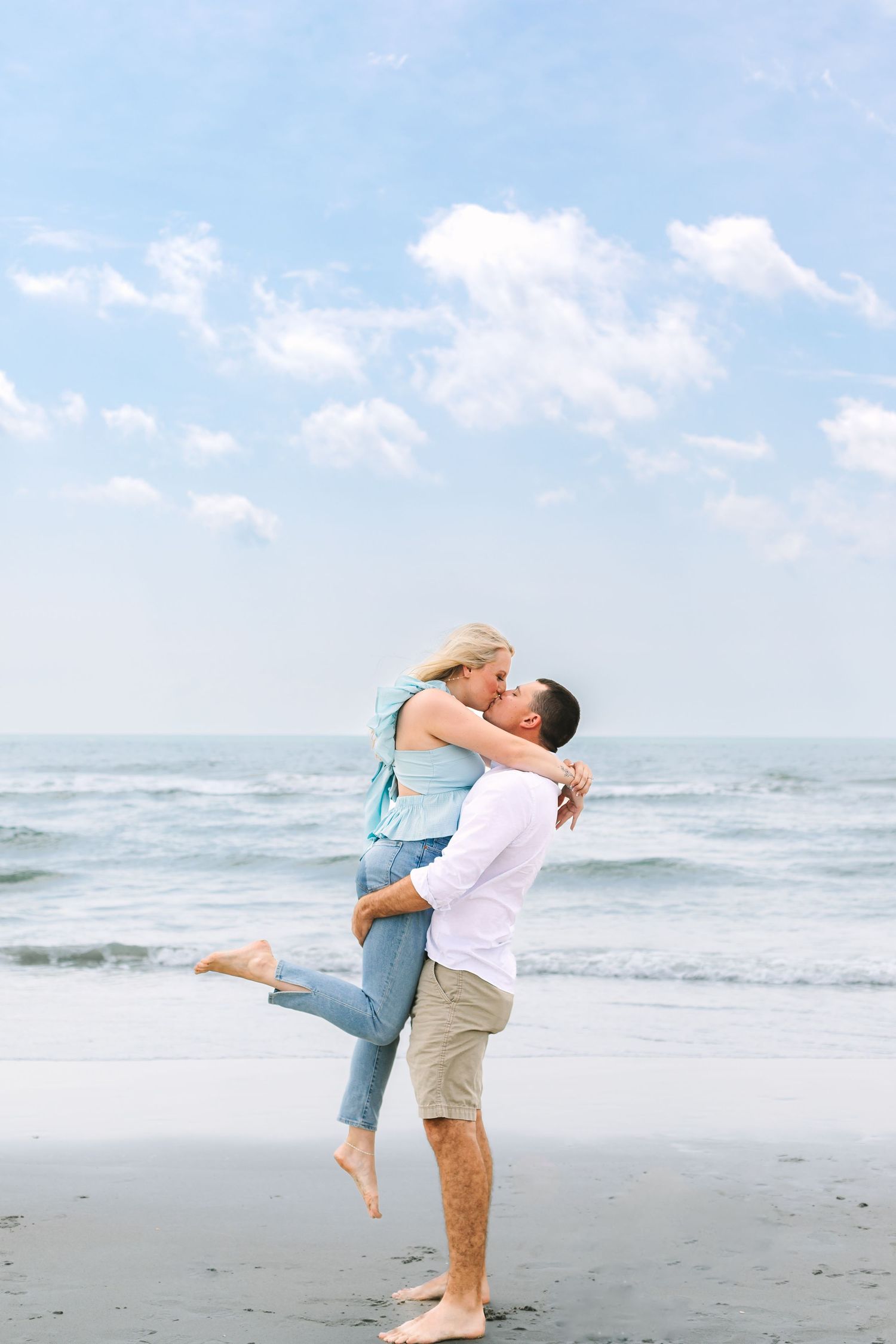 Man lifting and kissing woman on a sunny beach with blue sky and ocean waves behind them.