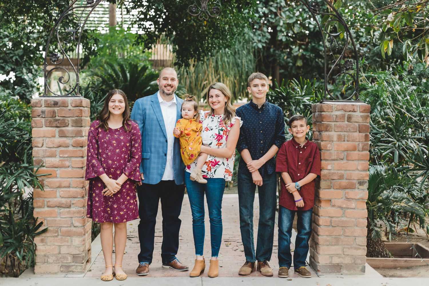 Family of six smiling together between brick pillars surrounded by lush tropical greenery.