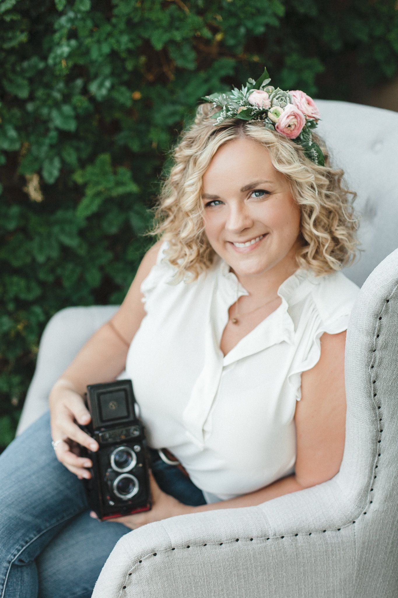 Smiling blonde photographer wearing a floral crown holding a vintage camera while seated outdoors.