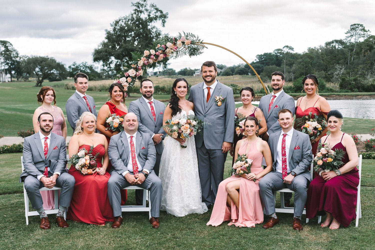 Wedding party group portrait with bride and groom in front of floral arch on a green golf course lawn.