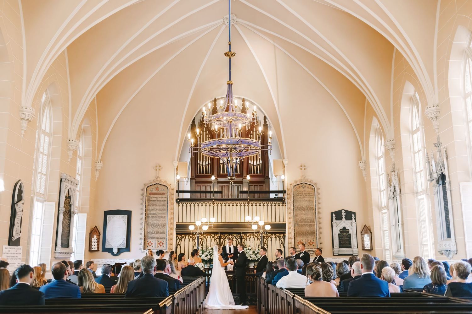 Wedding ceremony inside a beautiful historic church with vaulted ceilings, chandelier, and guests seated in wooden pews.