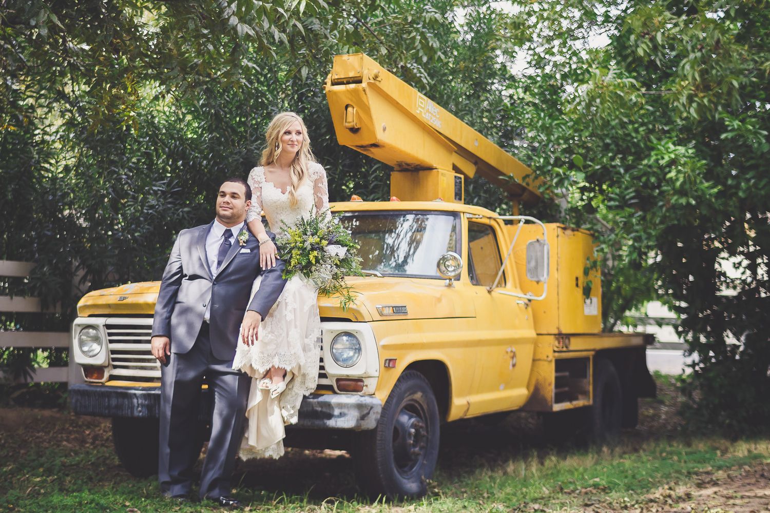 Bride and groom posing beside a vintage yellow utility truck surrounded by lush green trees.