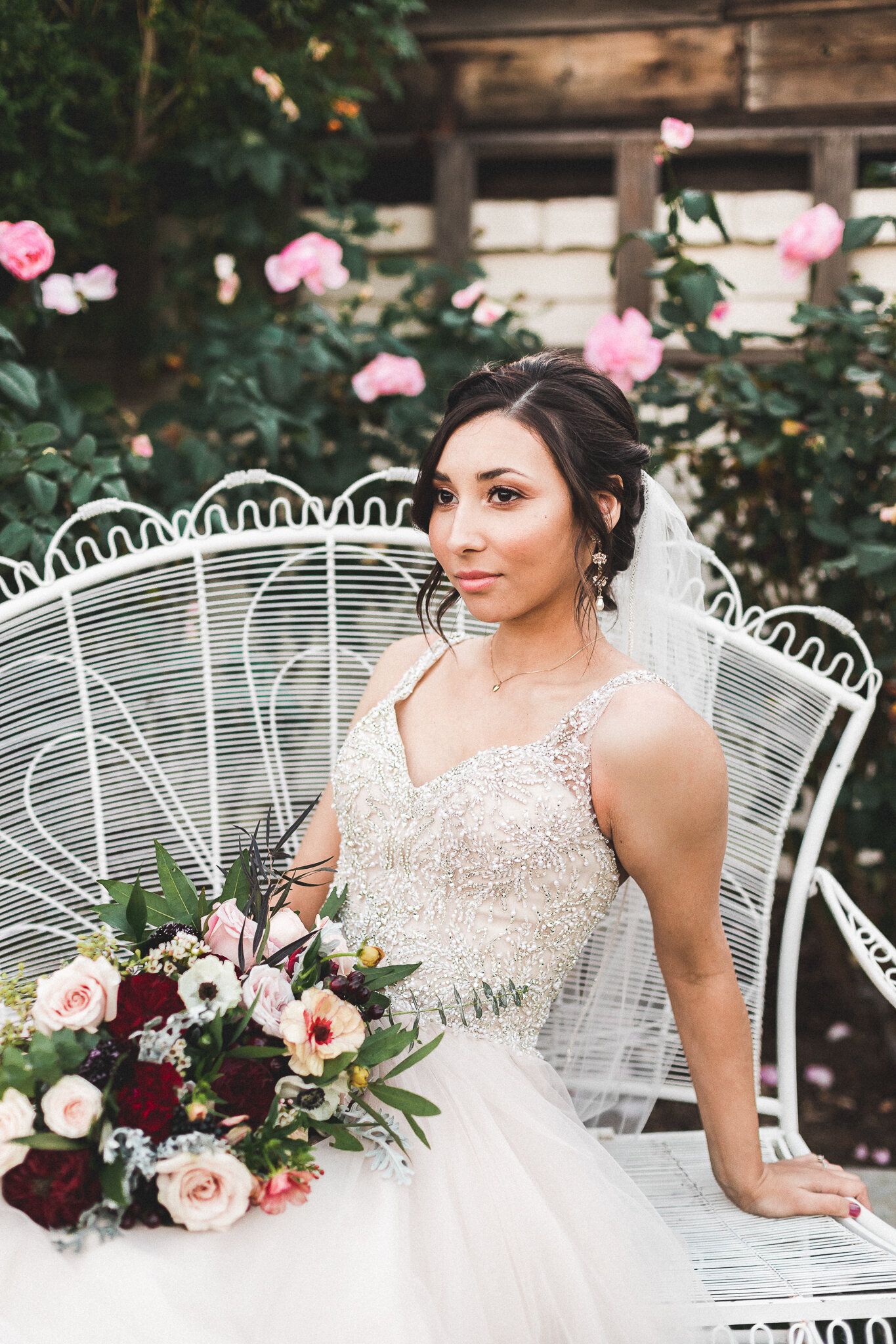 Elegant bride in beaded gown sitting on white iron chair surrounded by pink roses.