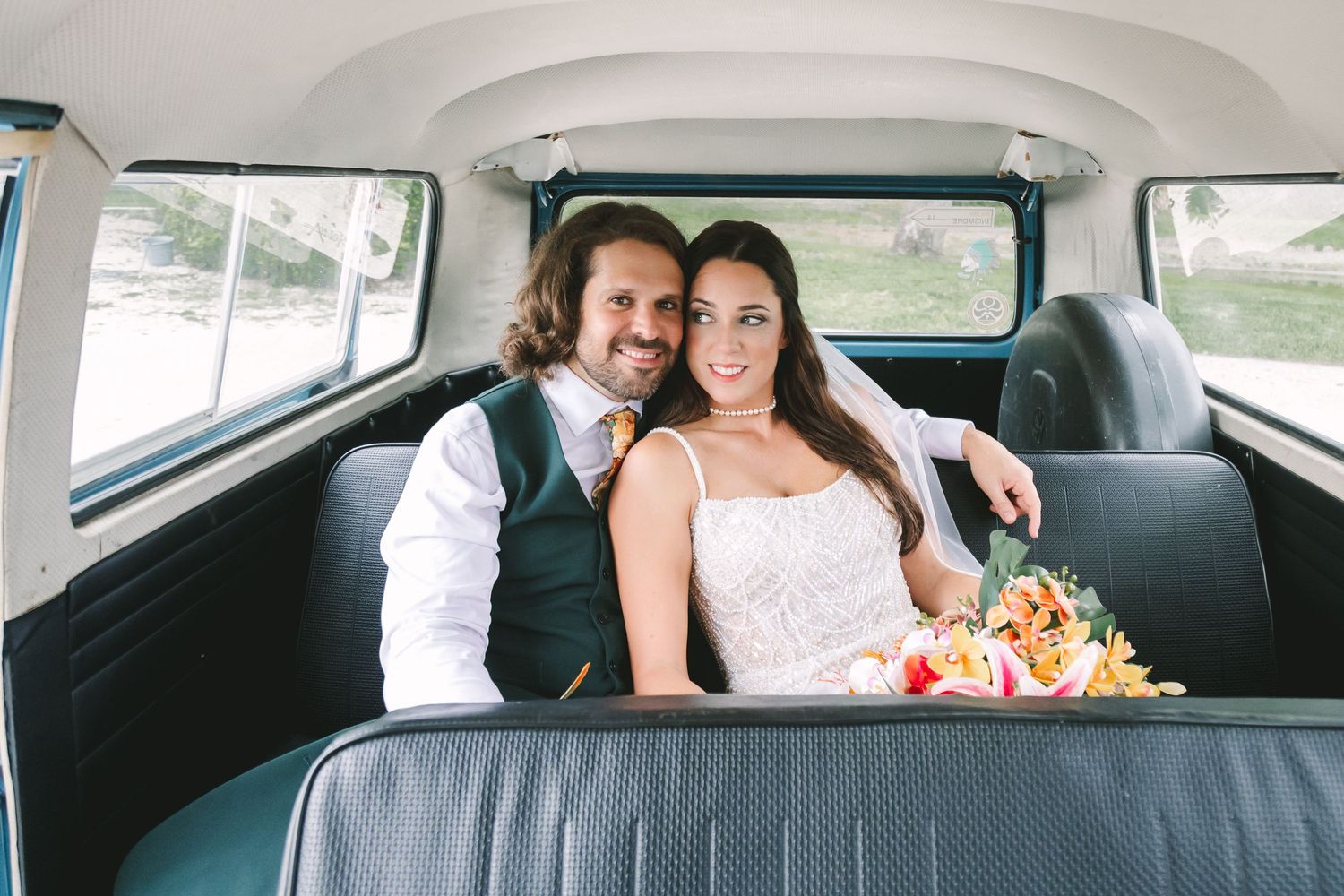 Bride and groom sitting together in the back seat of a vintage VW van, smiling with a colorful bouquet.
