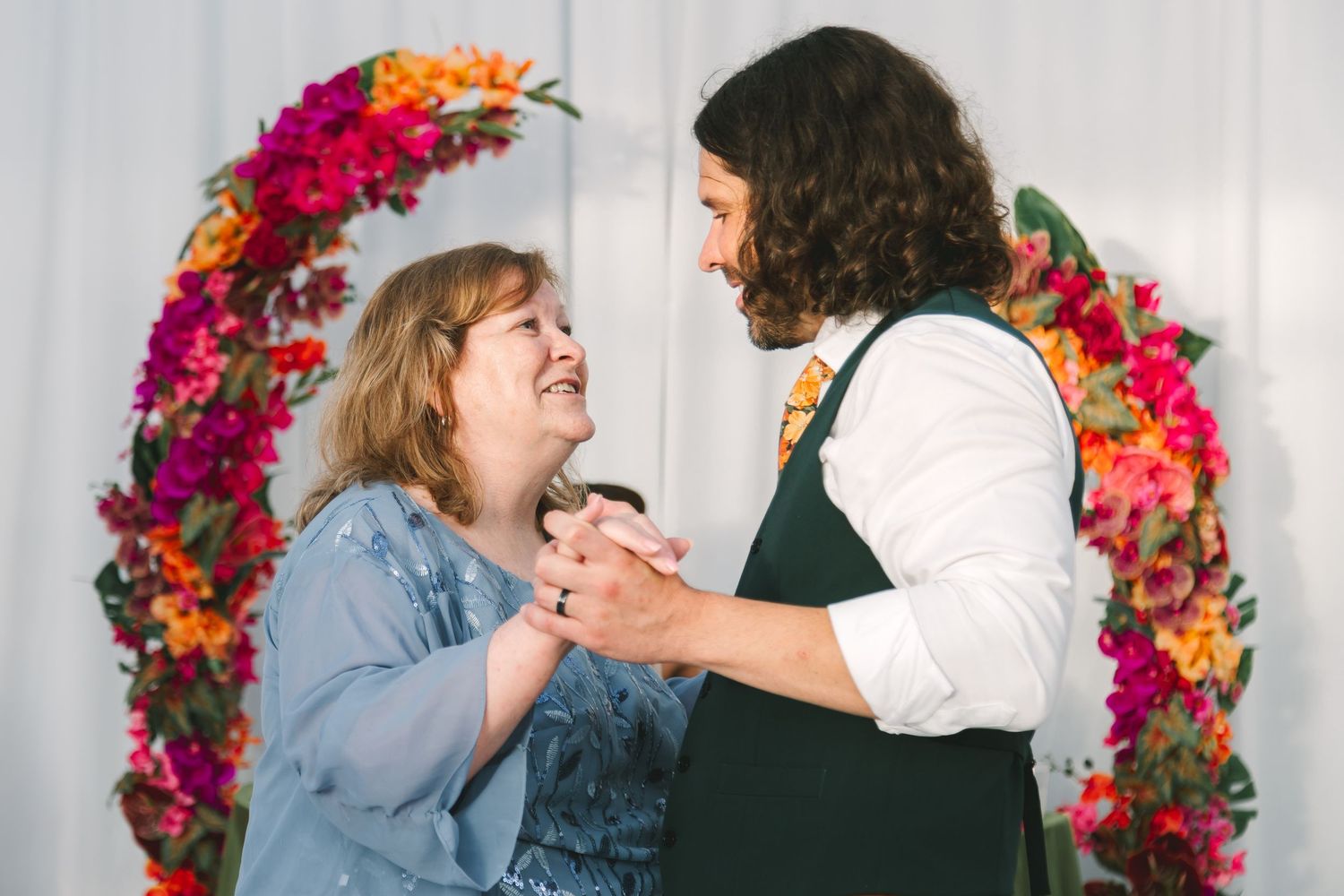 Bride and groom share a joyful dance in front of a vibrant floral arch at their wedding reception.