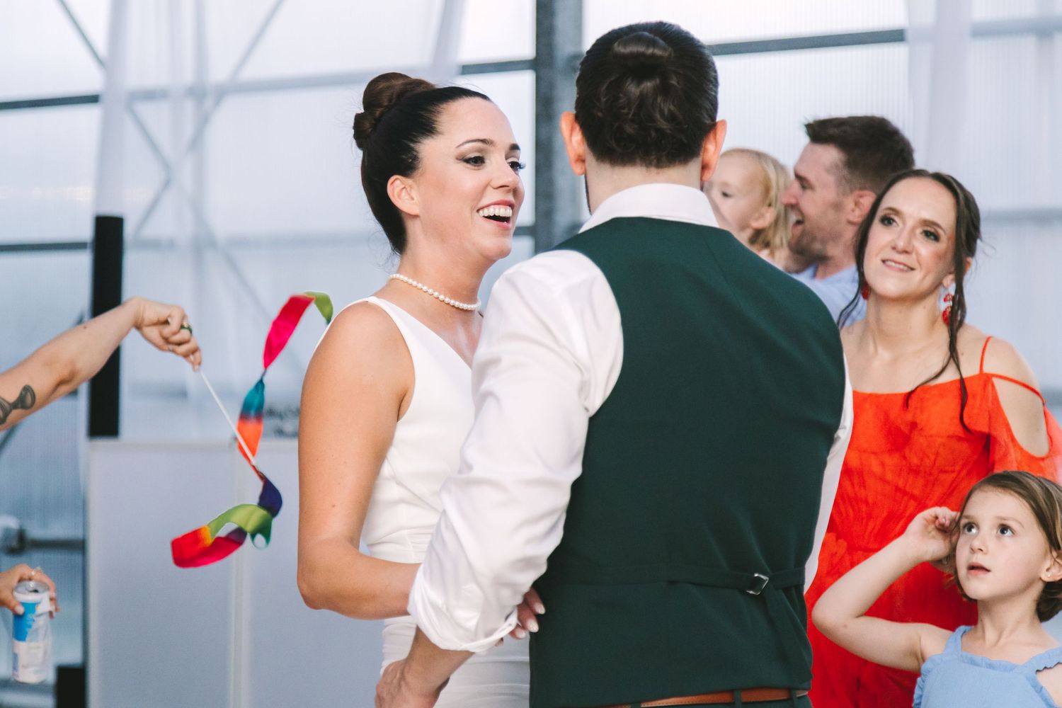 Bride in white dress laughs with groom in green vest while guests celebrate at wedding reception.