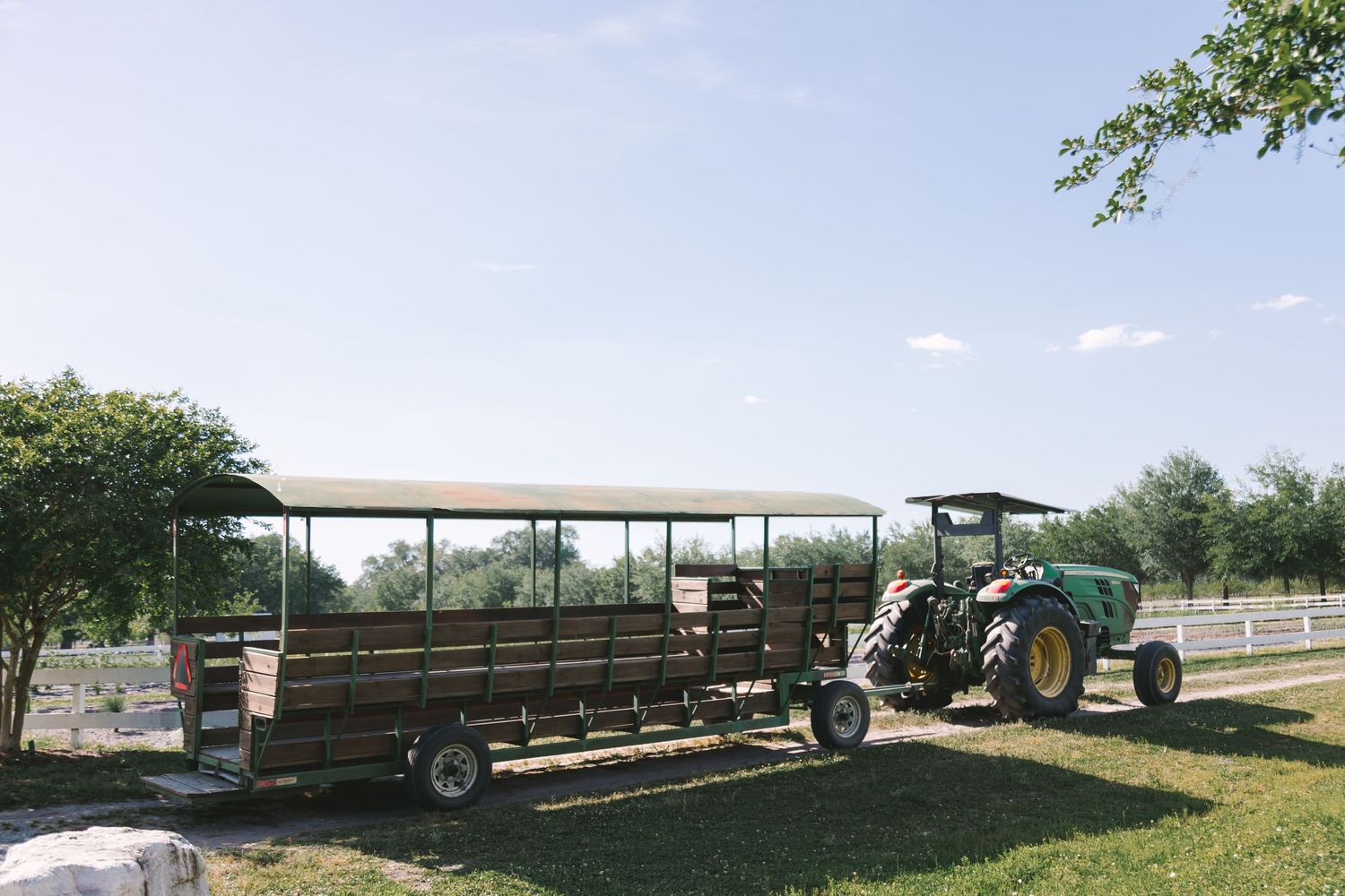 Green John Deere tractor pulling a covered wagon hayride trailer on a sunny farm with lush green trees.