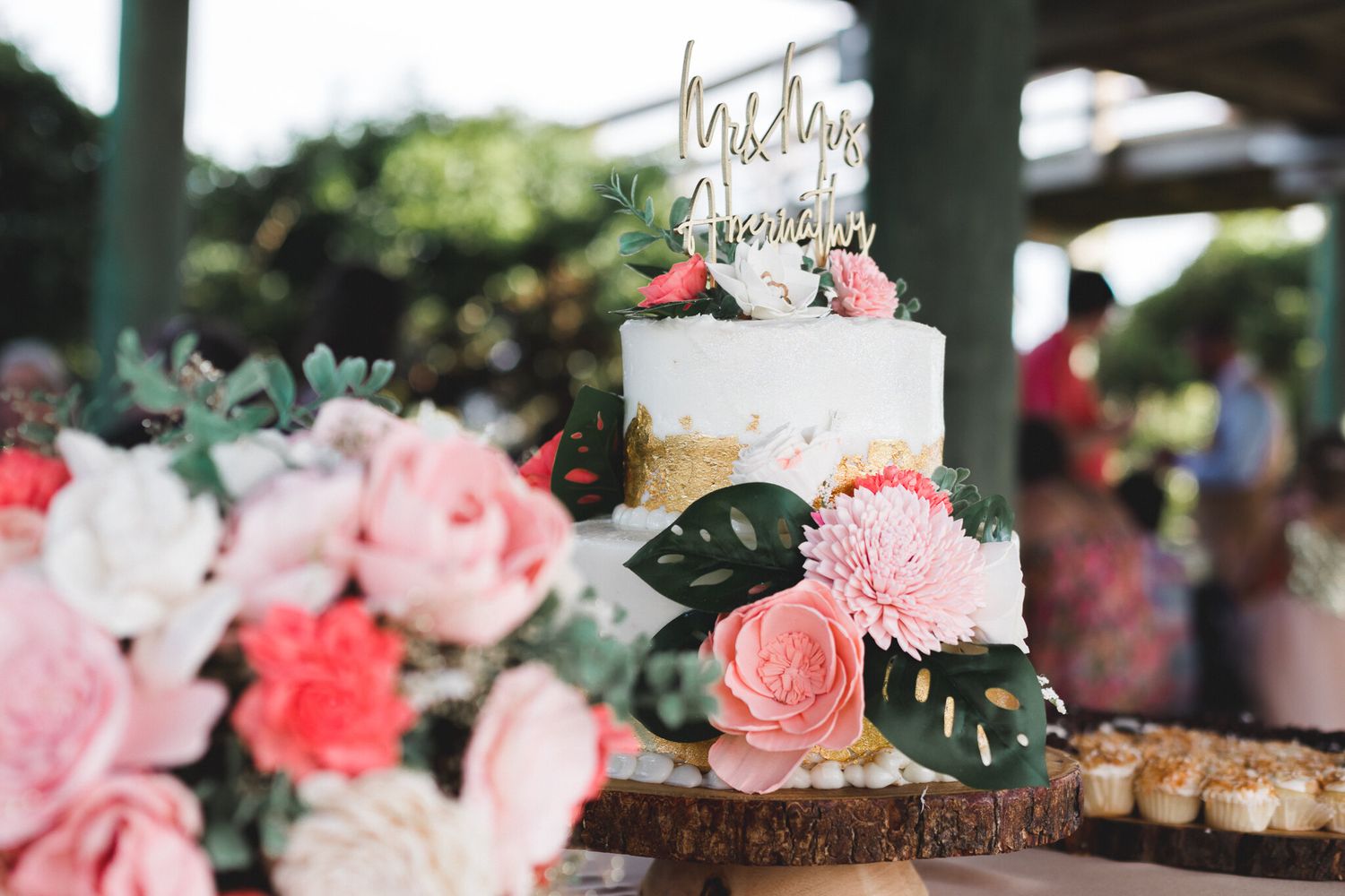 Two-tier white and gold wedding cake with pink floral decorations on rustic wood stand.
