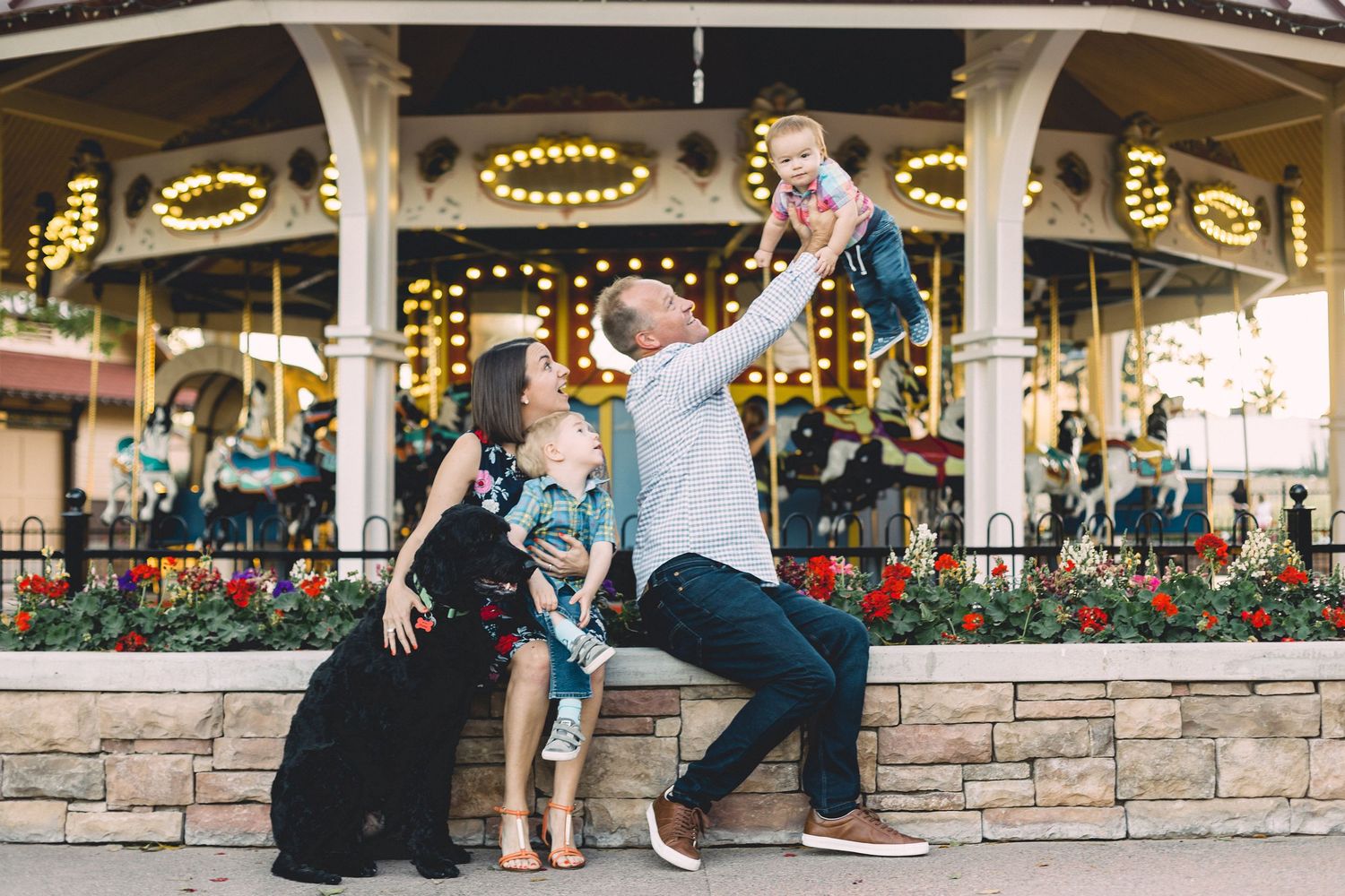 Happy family with a dog and baby posing playfully in front of a colorful carousel at dusk.
