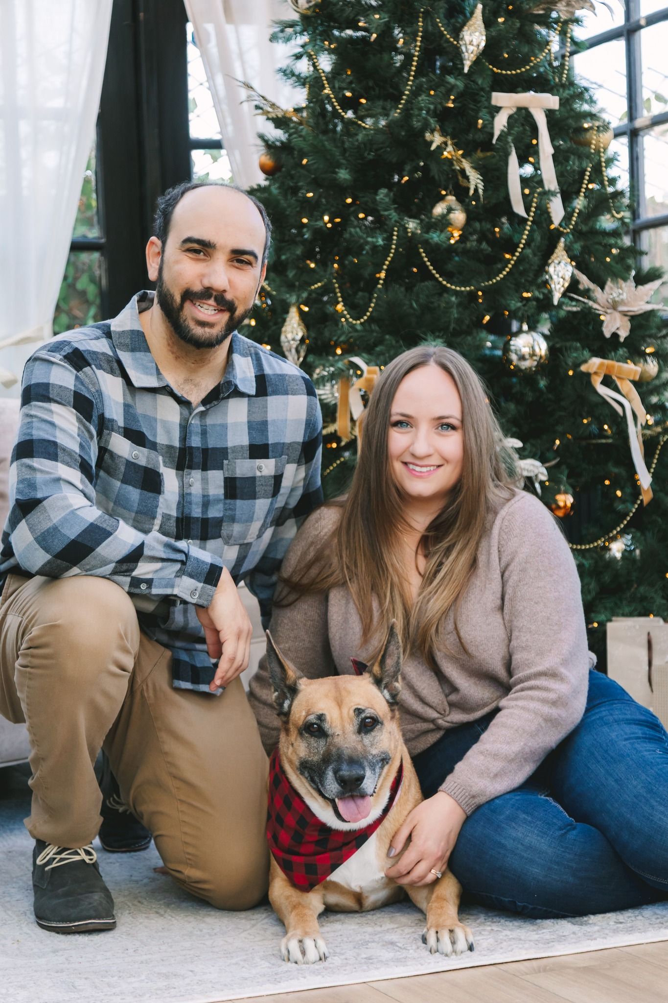Smiling couple posing with their dog wearing a plaid bandana in front of a decorated Christmas tree.