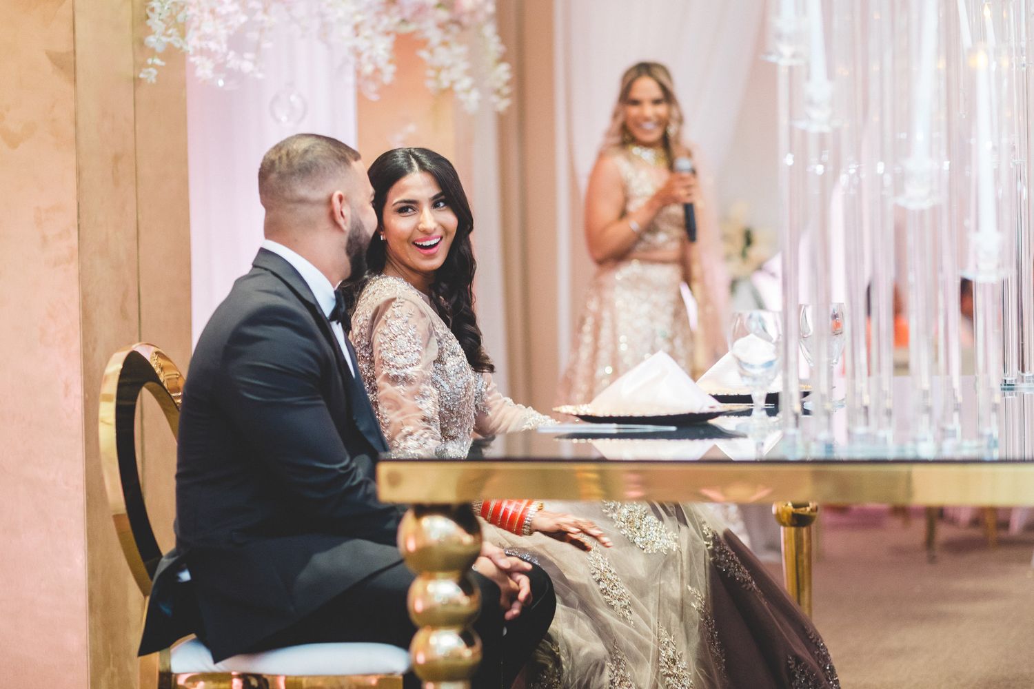 A smiling South Asian bride in a beige embroidered outfit sits with her groom at a beautifully decorated wedding reception.