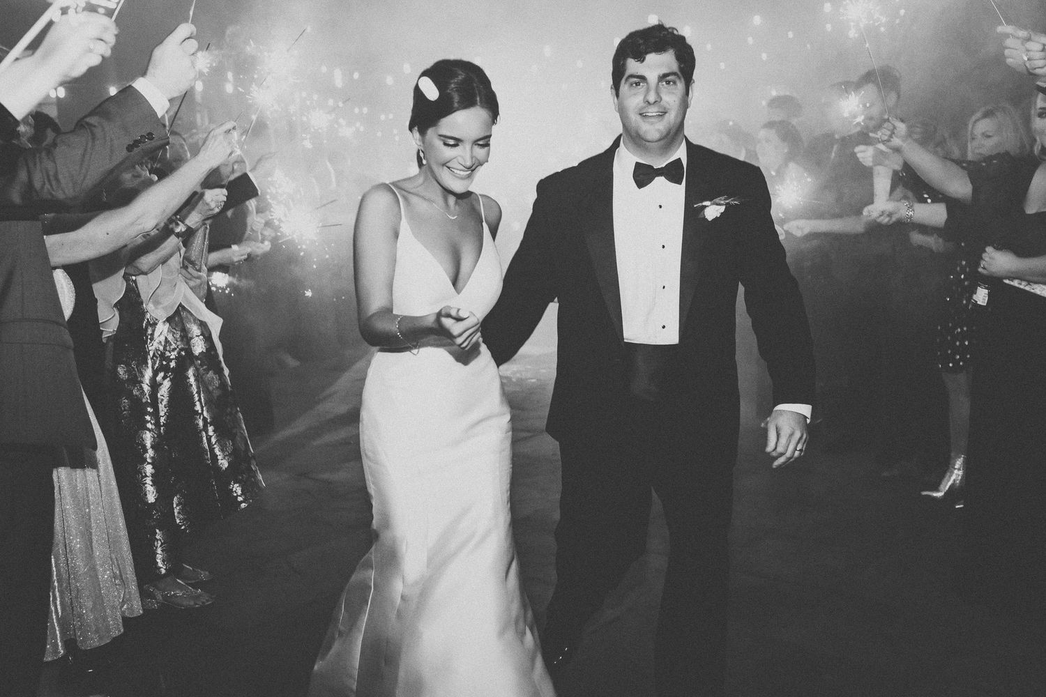 Black and white photo of bride and groom walking through sparkler exit surrounded by cheering wedding guests.