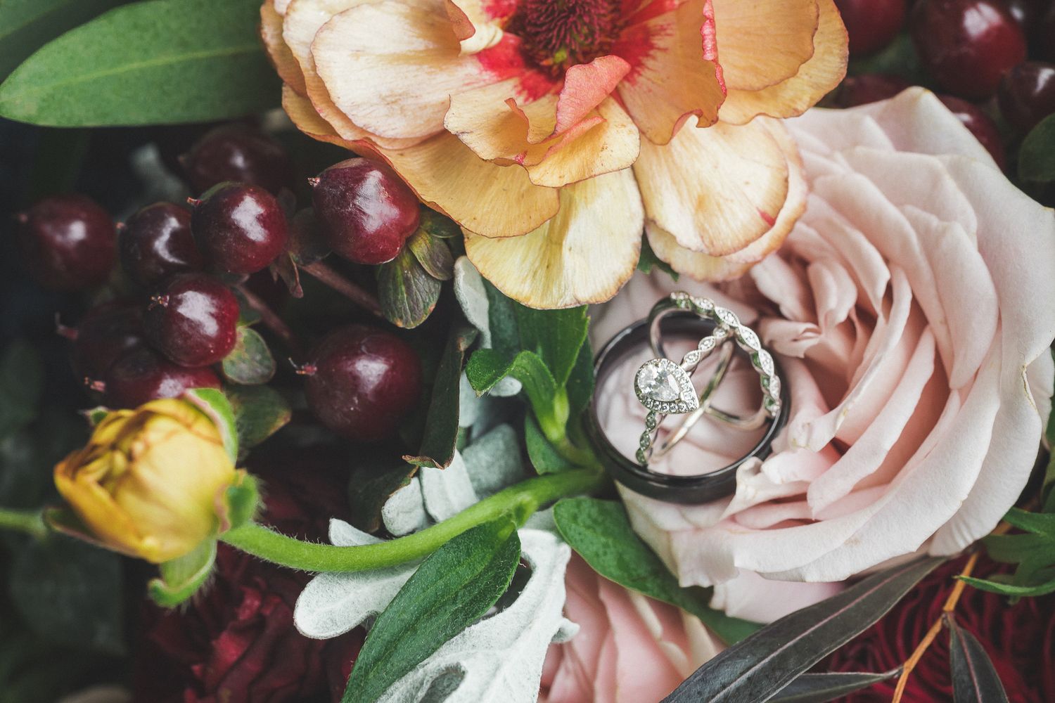Wedding rings resting on a bridal bouquet with roses, ranunculus, and fresh cherries.