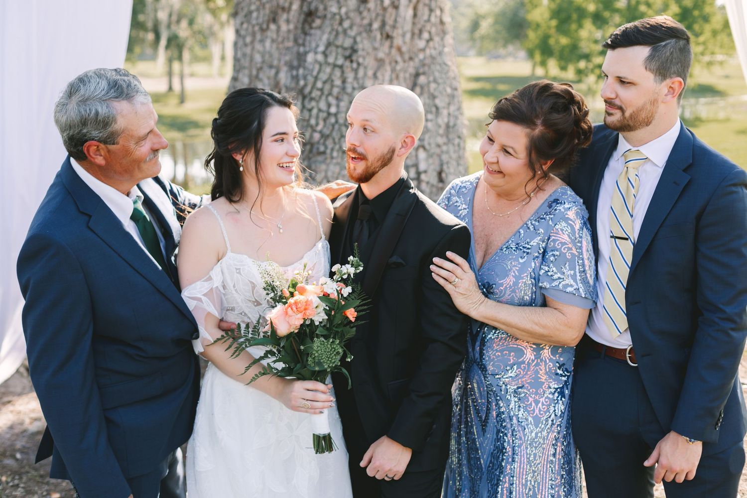 A bride and groom pose with family members in formal attire outdoors near a tree at a wedding celebration.