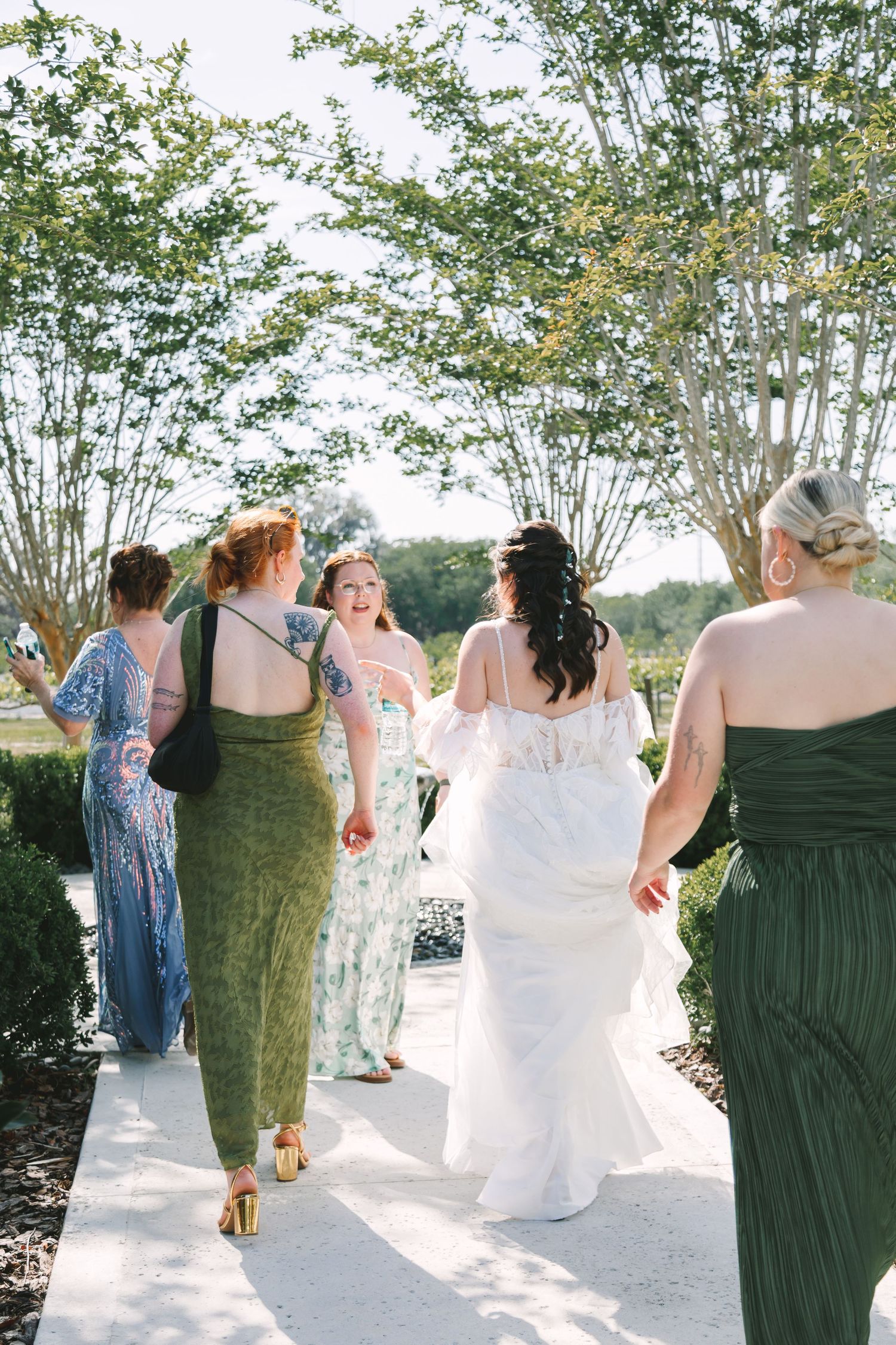 Bride in white gown walks with bridesmaids in green dresses along sunny outdoor pathway.