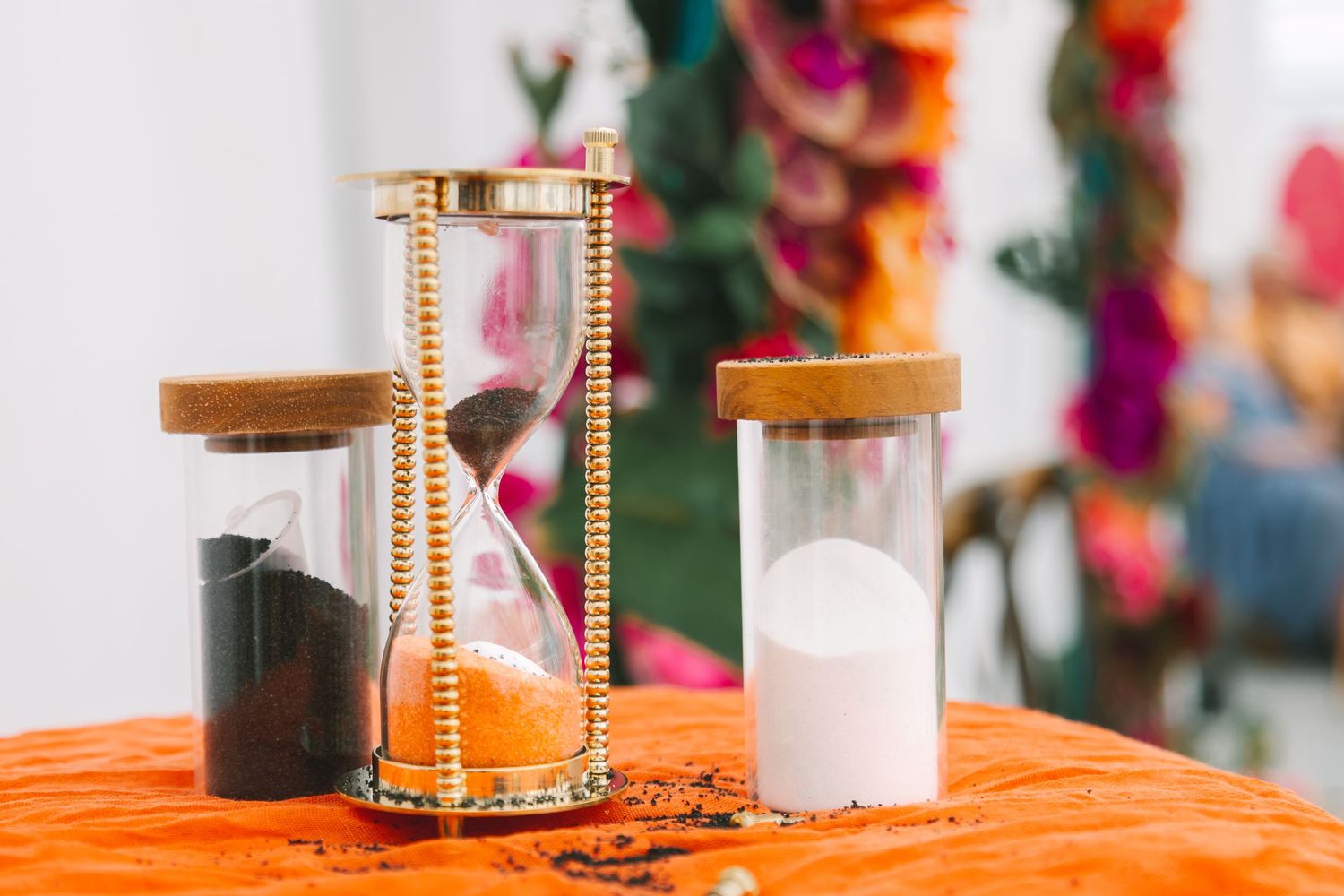Three decorative hourglasses with black, orange, and white sand displayed on an orange tablecloth at a colorful event.