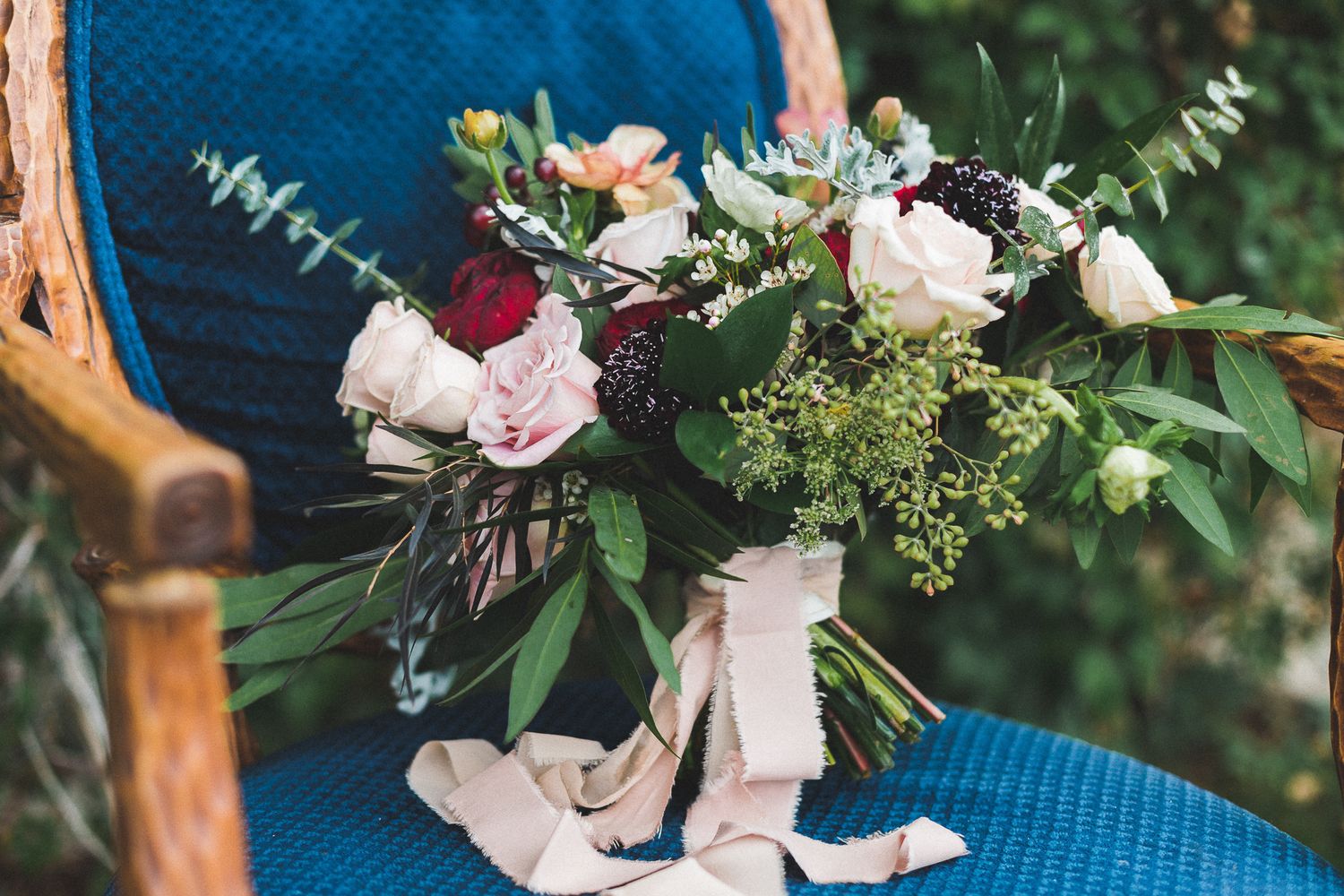 Lush bridal bouquet with burgundy and white roses, greenery, and blush ribbon on blue chair.