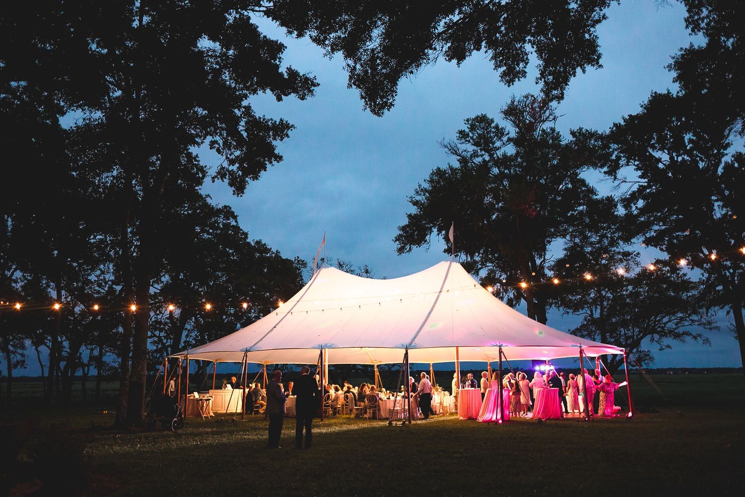 Illuminated white wedding tent glowing at dusk surrounded by trees with string lights.