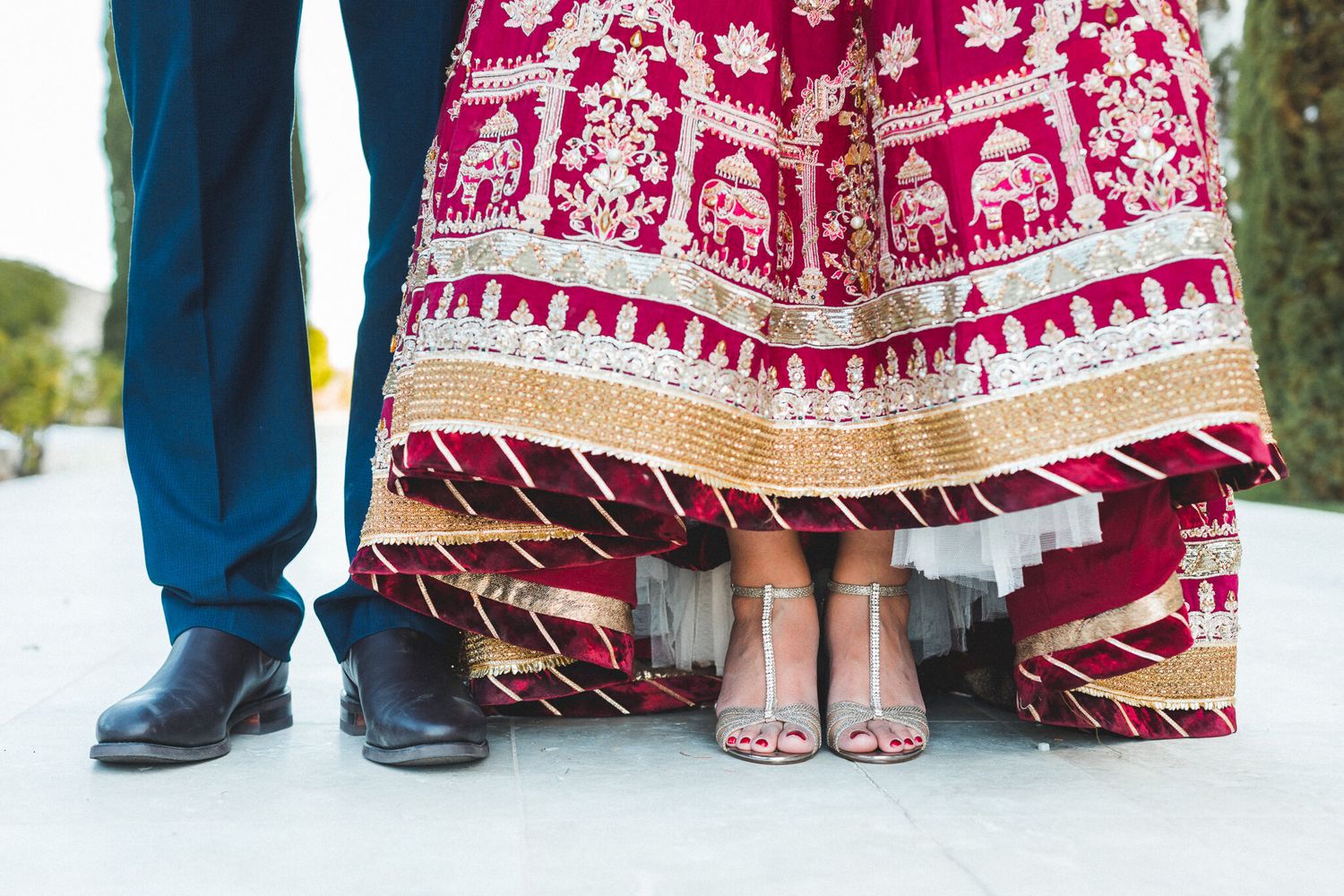 Close-up of South Asian bride in red embroidered lehenga hemline with silver heels and groom shoes.