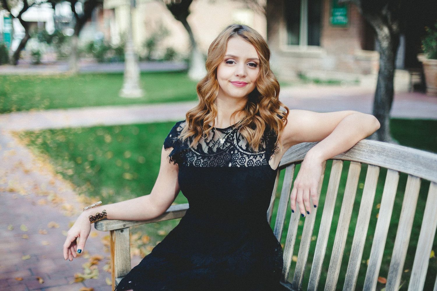 Young woman with wavy blonde hair posing on wooden bench in elegant black lace dress.