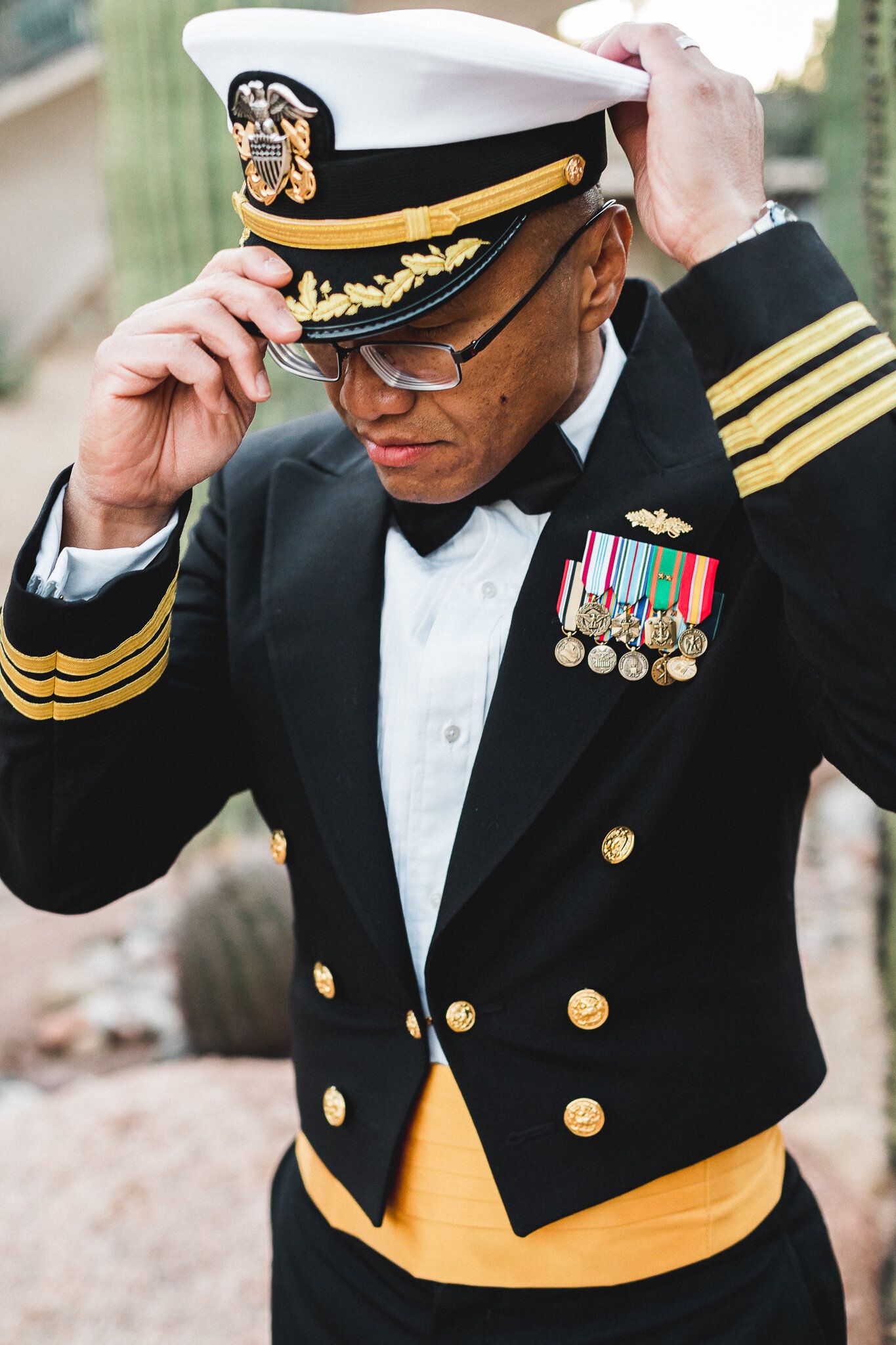 Distinguished military officer in formal Navy dress uniform adjusting white cap outdoors.