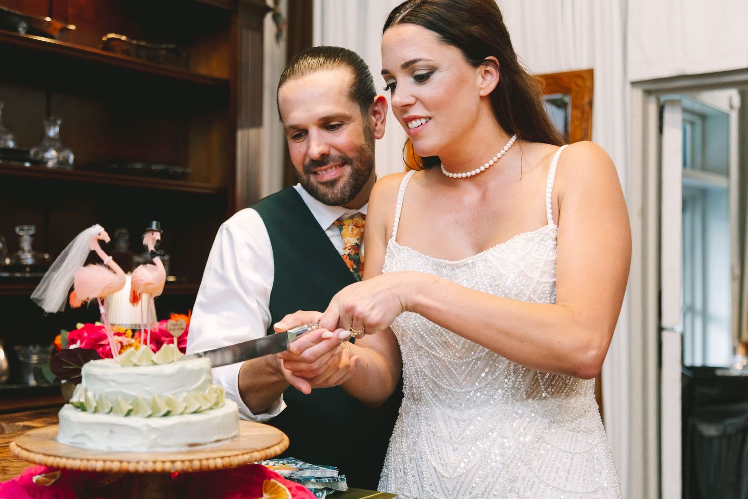 Bride and groom smiling while cutting white wedding cake at reception.