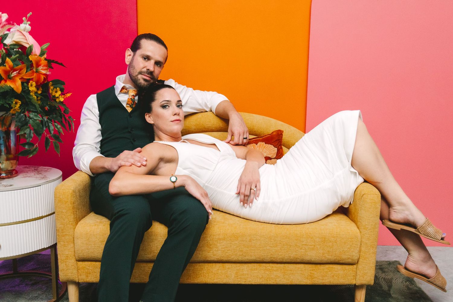 Couple posing on yellow sofa against colorful pink and orange wall, woman in white dress.