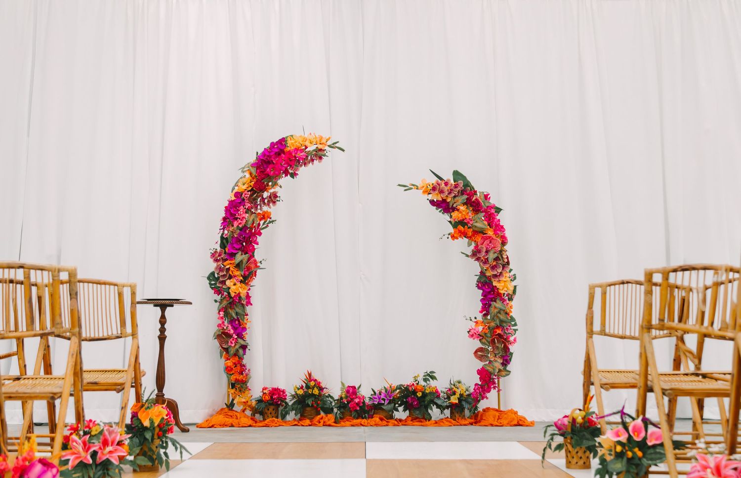Vibrant floral arch with pink, orange, and purple blooms frames a wedding ceremony altar against white drape backdrop.