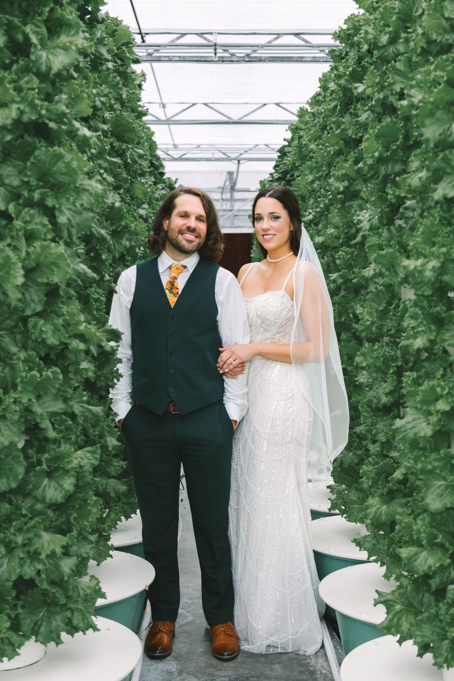 Bride and groom holding hands smiling in lush green hydroponic greenhouse rows.