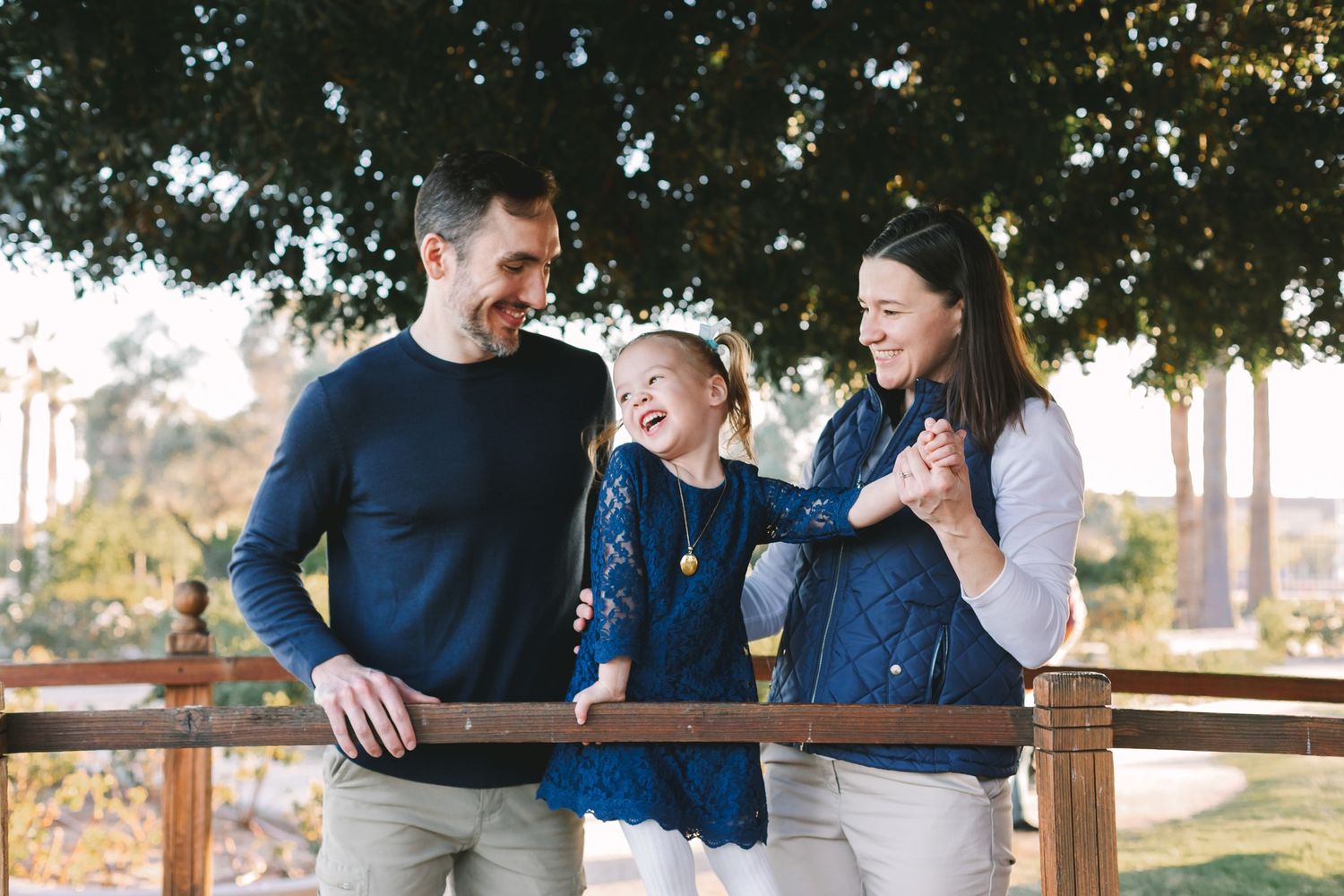 Laughing family of three leaning on wooden fence rail under large tree outdoors.