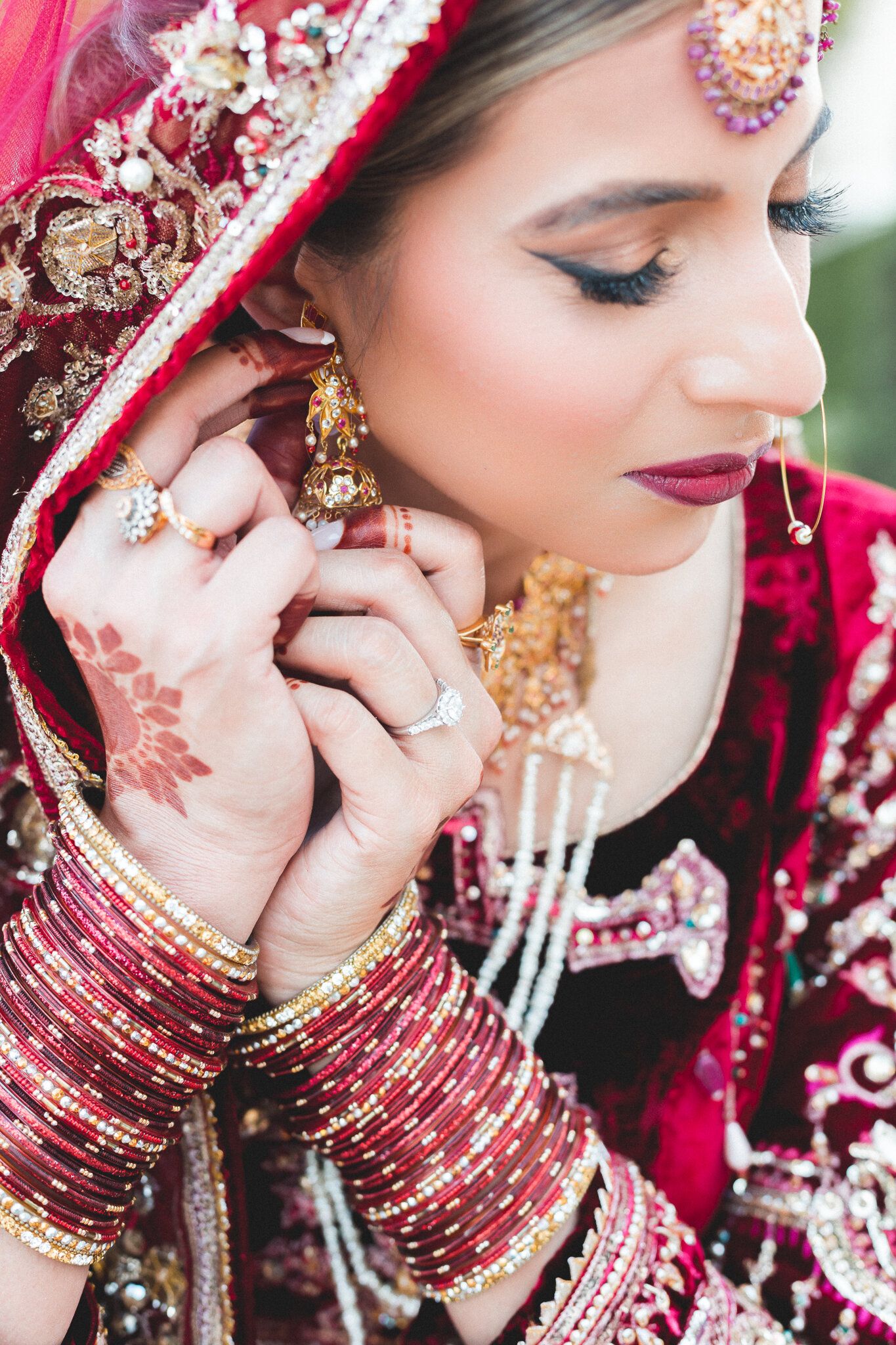 South Asian bride in red lehenga adjusting gold jhumka earrings wearing bangles and mehndi hands.