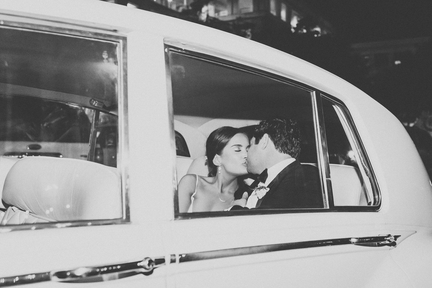 Black and white photo of newlyweds sharing a kiss inside a classic vintage wedding car at night.